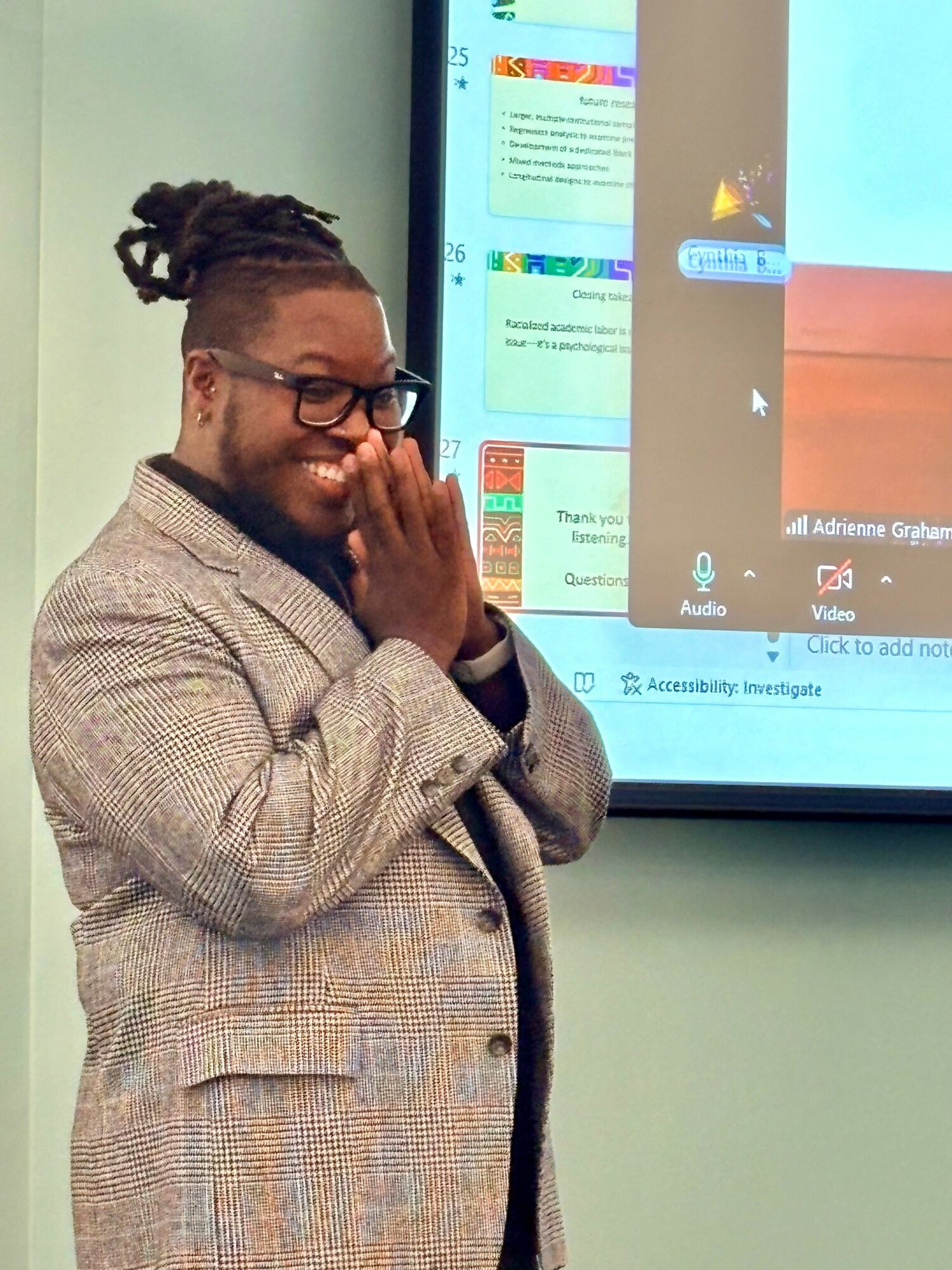 Person with glasses and dreadlocks, wearing a blazer, smiling and covering mouth with hand, standing in front of a screen.