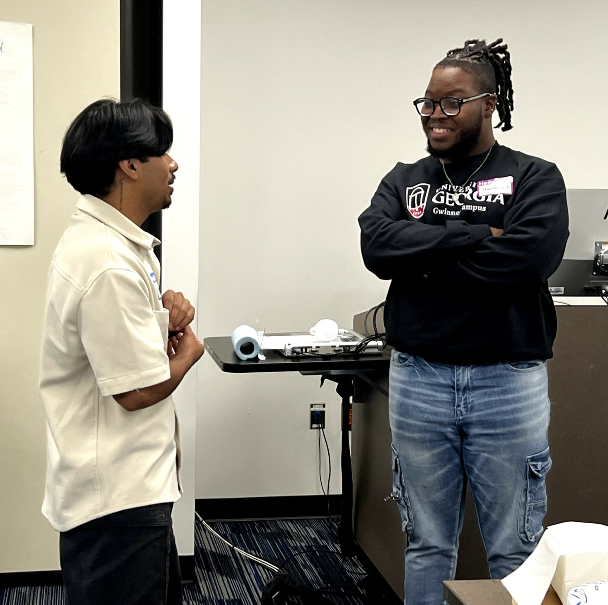 Two people talking indoors, one with short black hair and the other with glasses and dreadlocks, smiling and engaging in conversation.