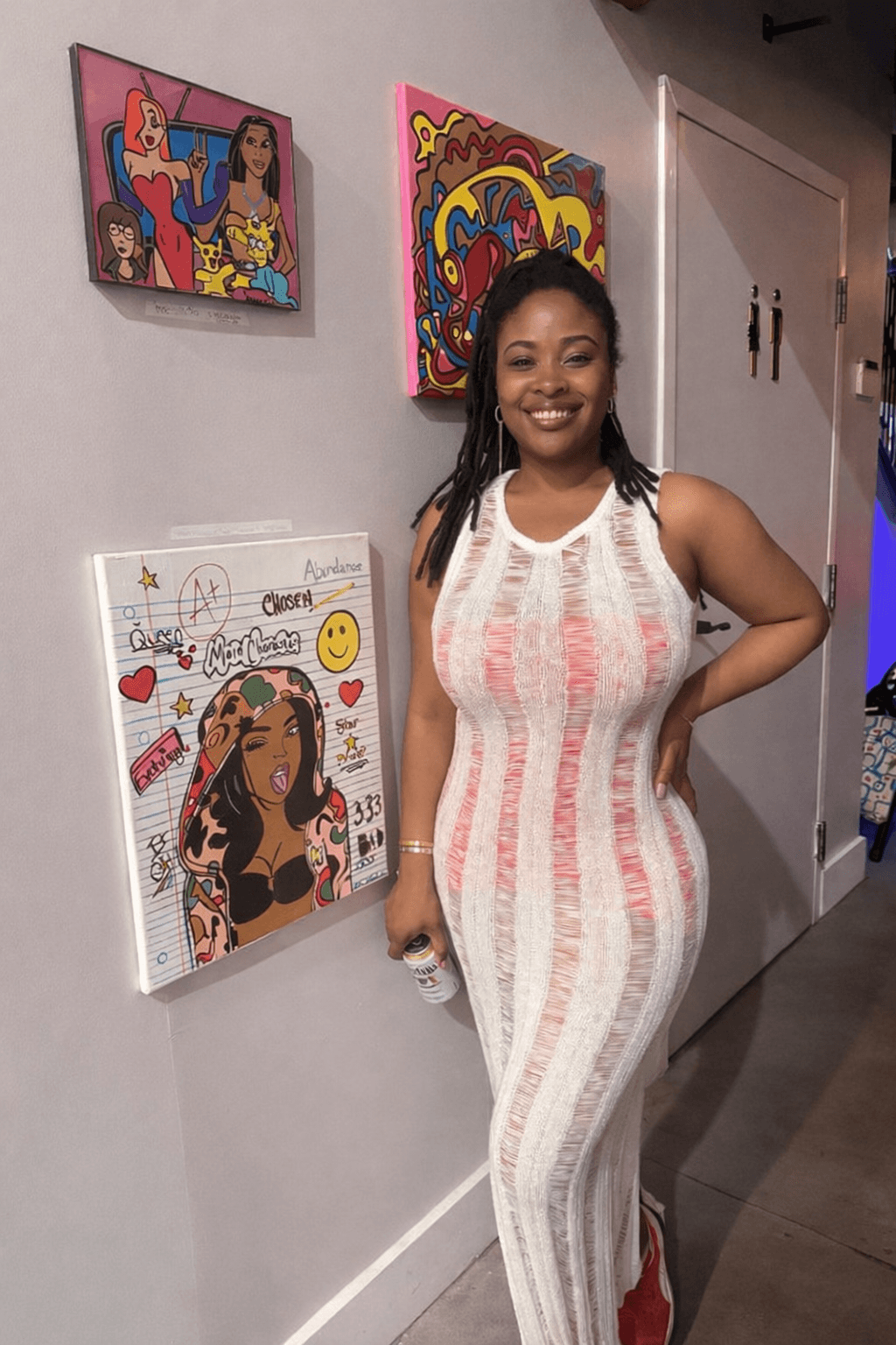 Smiling woman in a striped sleeveless jumpsuit standing indoors near colorful artwork on the wall.