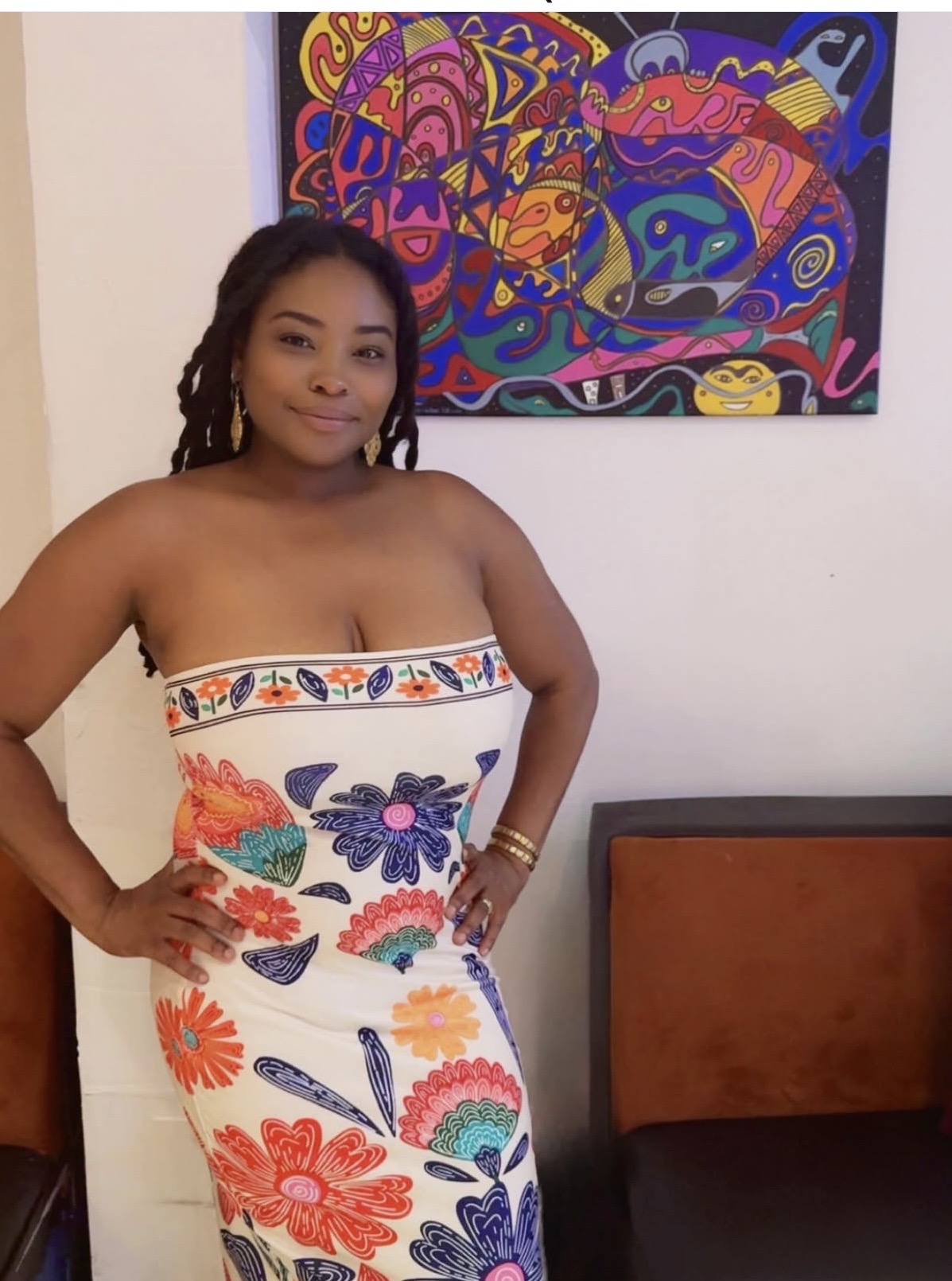 Woman in a strapless dress with colorful floral and butterfly patterns, standing indoors with artwork behind her.