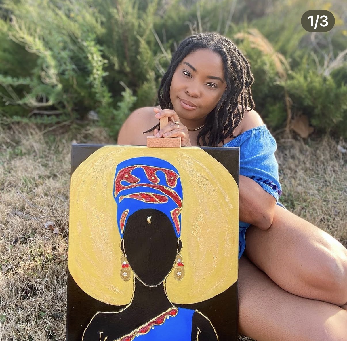 Young woman sitting outdoors with a colorful painting of a woman in traditional attire, holding her chin, smiling.