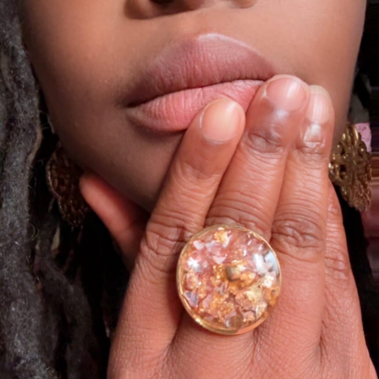 Close-up of a person's lips and hand with a large, colorful ring, fingers touching lips, skin tone visible.
