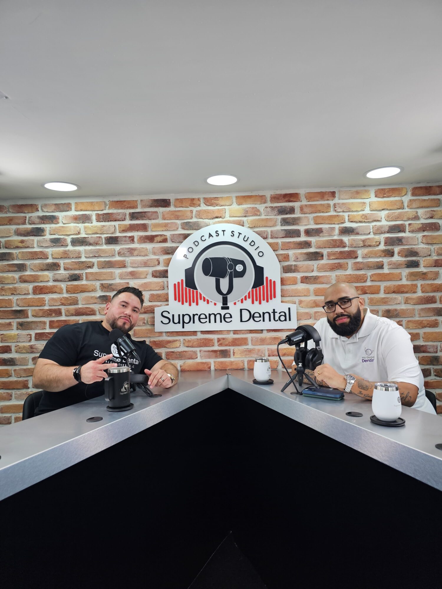 Two men sit at a table in a dental studio with a brick wall background and a sign reading 'Supreme Dental'.