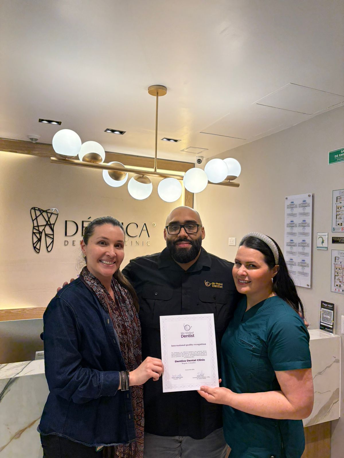 Three people standing indoors, holding a certificate, smiling, with a modern light fixture above and framed documents on the wall.