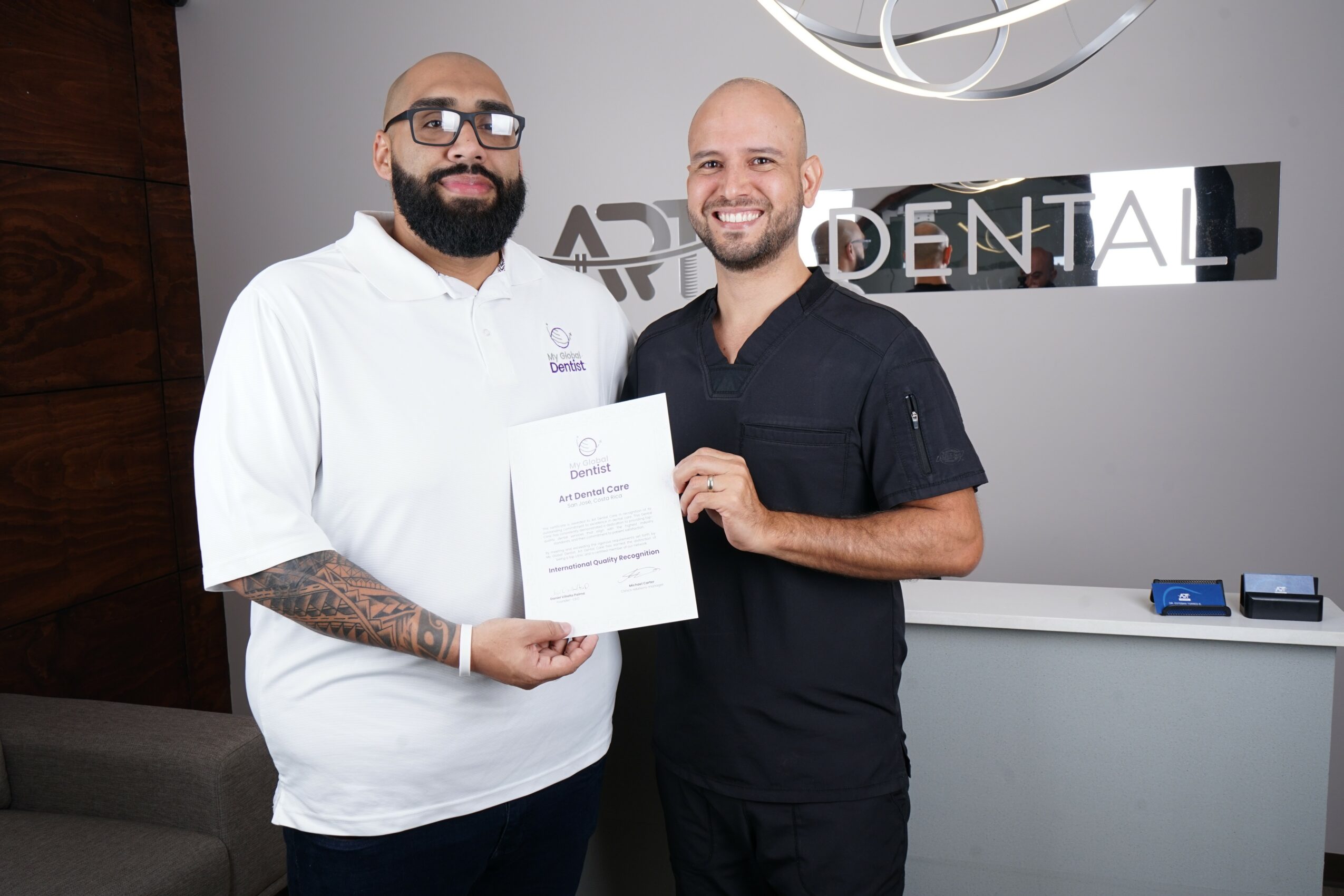 Two men standing indoors, one holding a certificate, smiling, with a sign reading 'DENTAL' behind them.