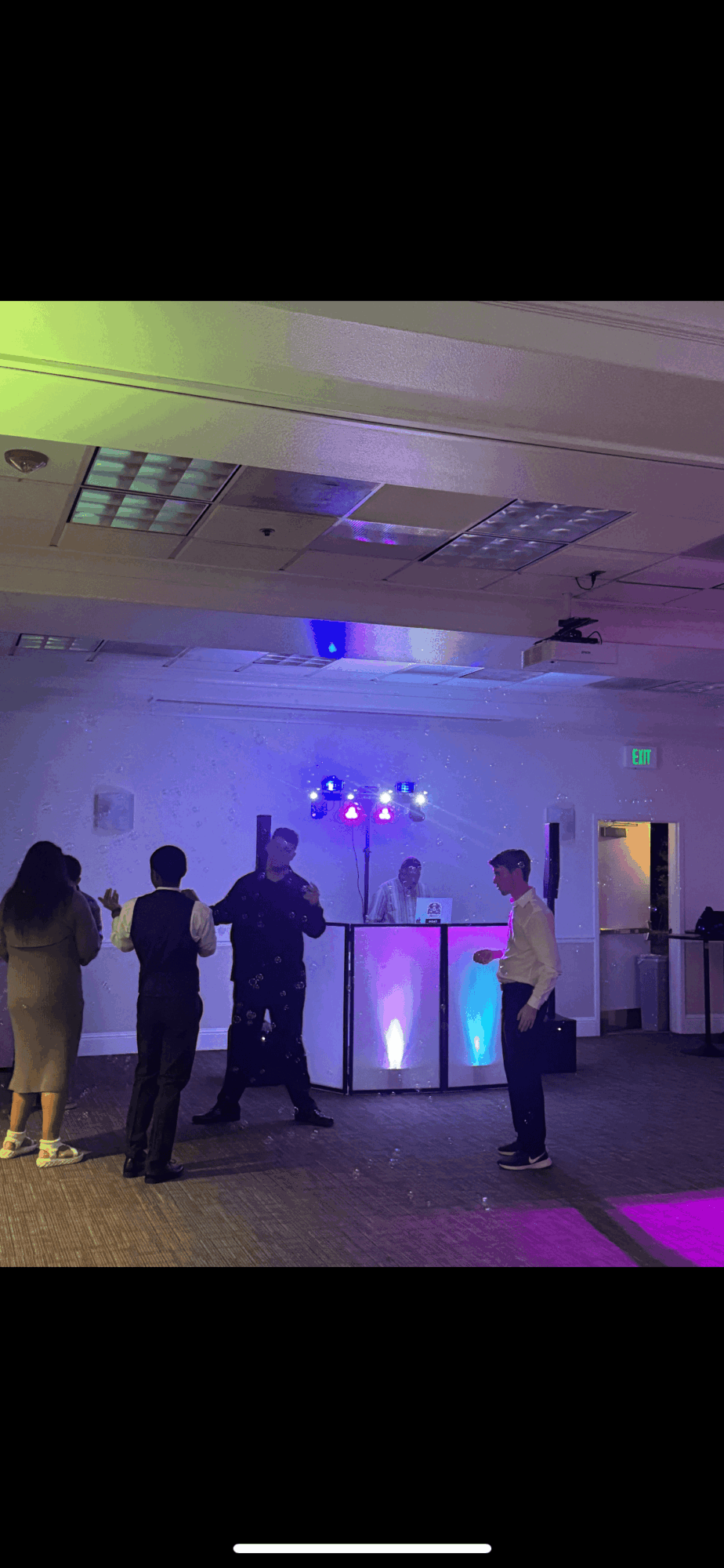 People gathered around a DJ booth with colorful lights in a large indoor space.