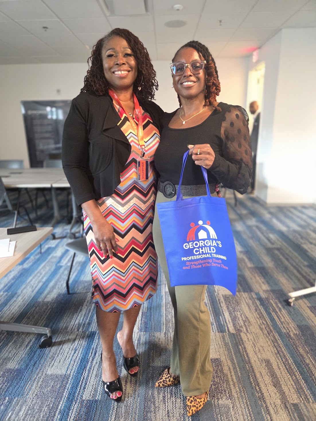 Two women standing indoors, smiling, one holding a blue bag with a logo, both dressed professionally.