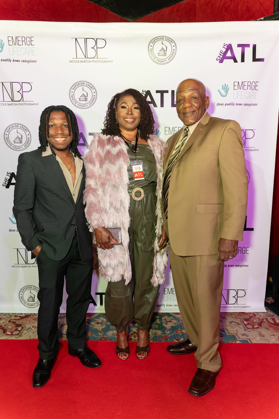 Three people standing on a red carpet in front of a backdrop with logos, smiling for the photo.