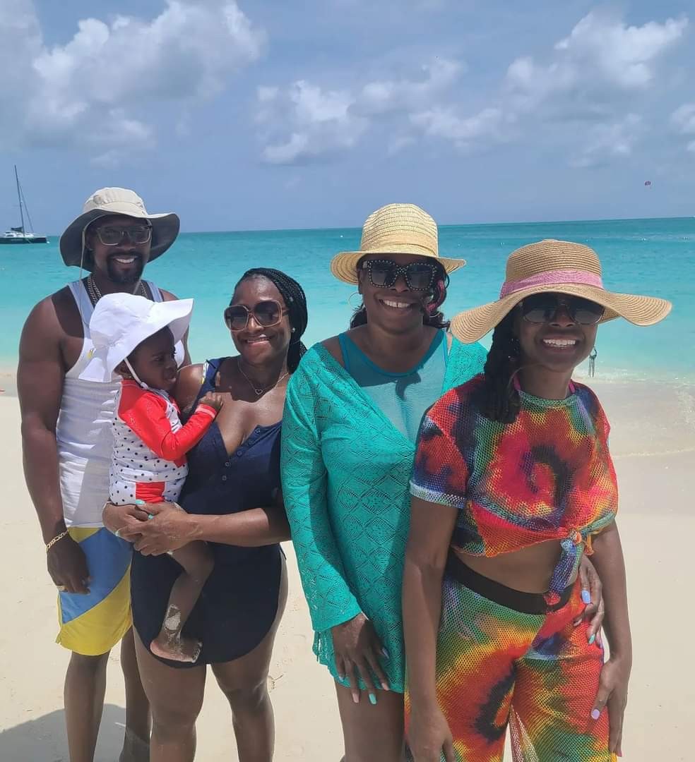 Four people, including a child, stand on a beach with ocean and sky in background, smiling and wearing summer clothes and hats.