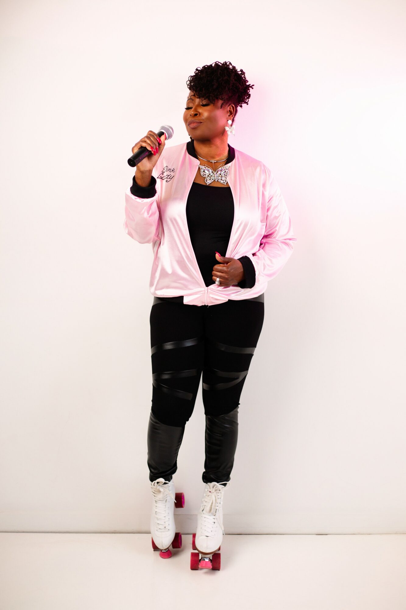 Person standing on roller skates holding a microphone, wearing a pink jacket, black pants, and a necklace, against a plain white background.