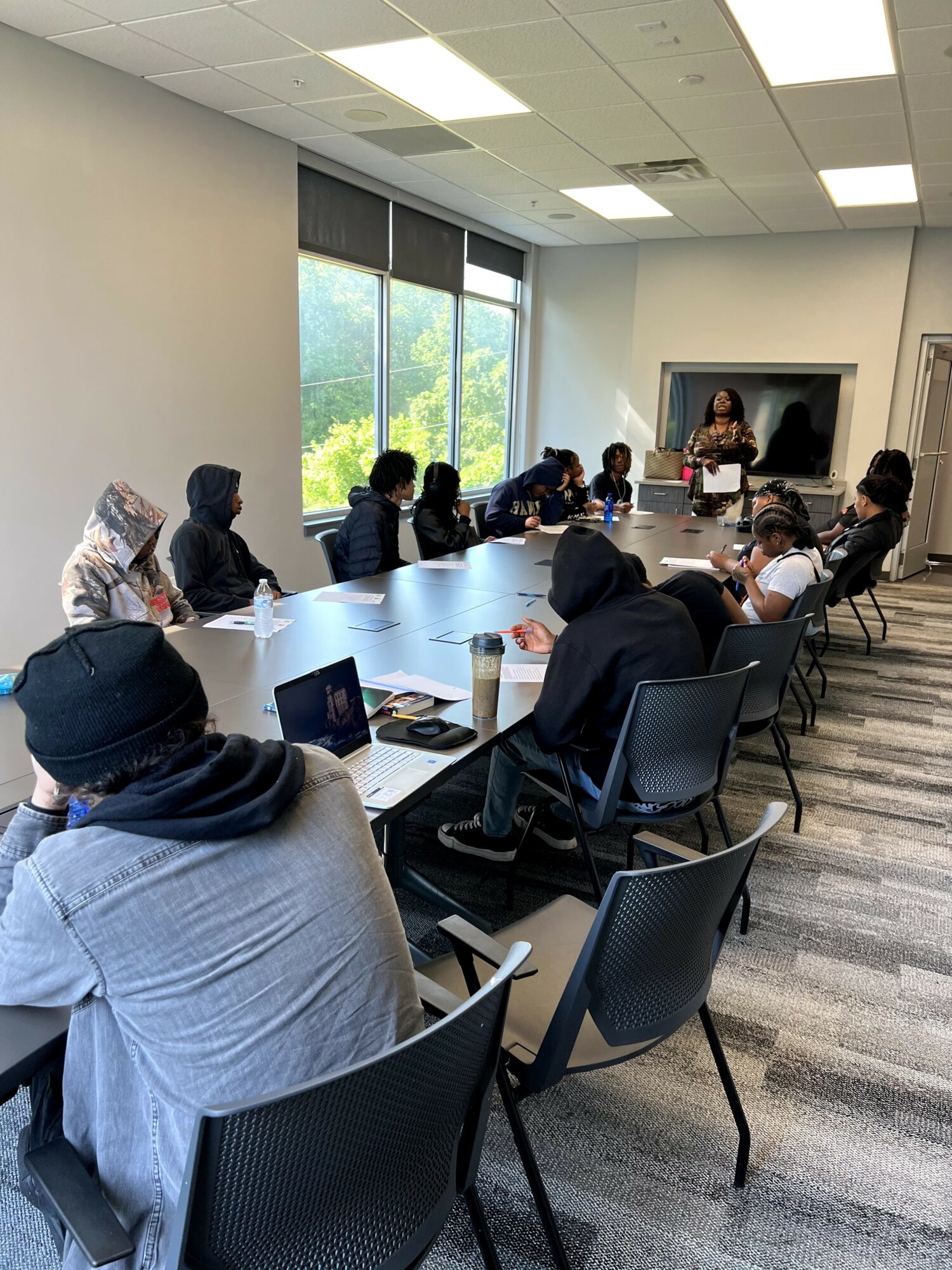 People seated around a conference table listening to a woman presenting, with large windows showing trees outside.