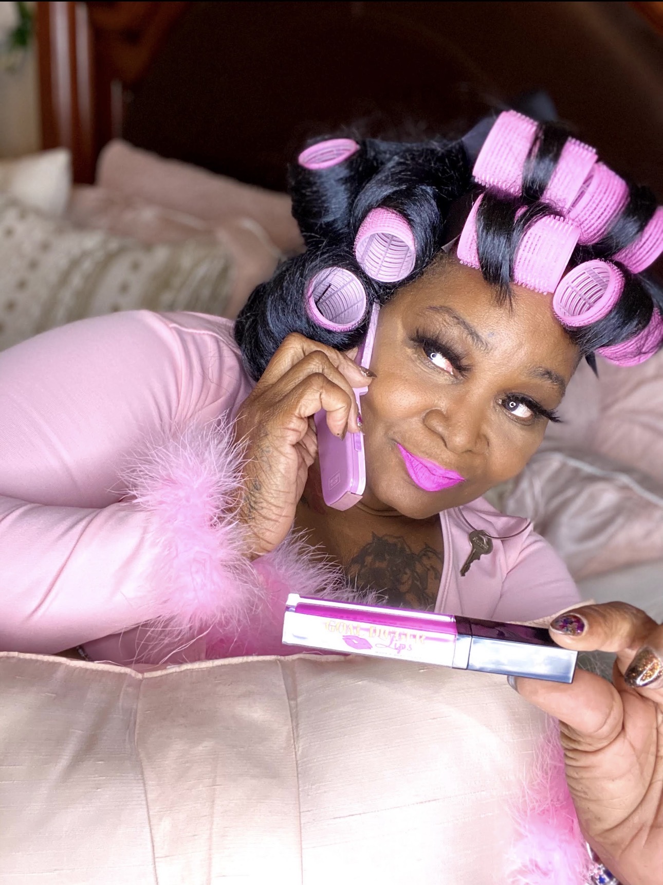Woman with pink lipstick and curlers in hair, lying on bed, talking on pink phone, wearing pink fuzzy robe.