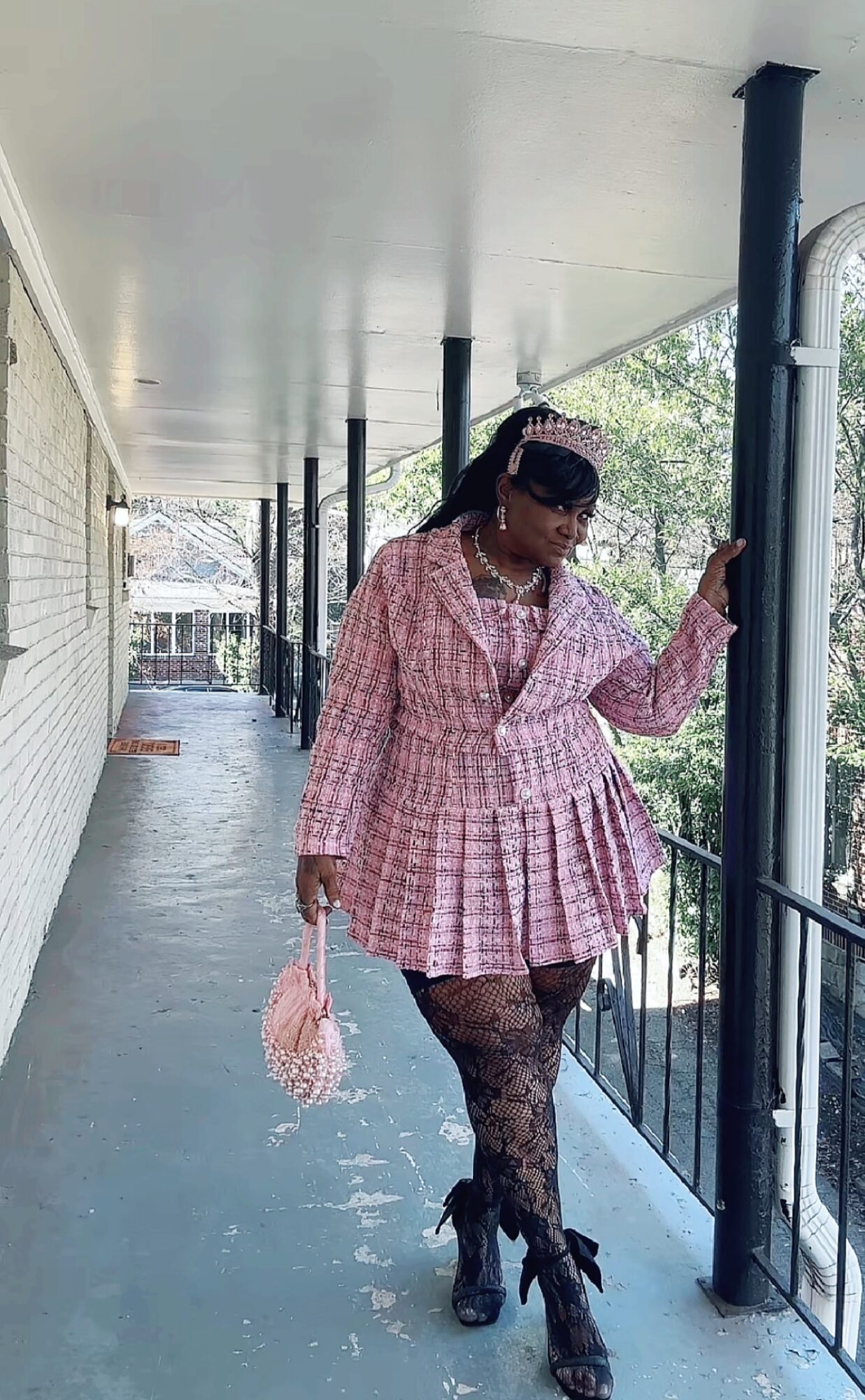 Person standing on a balcony, wearing a pink checkered outfit, holding a pink bag, with trees and buildings in the background.