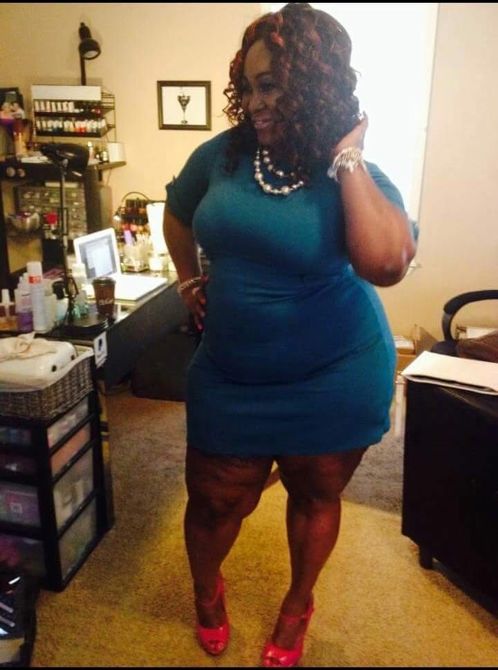 Woman in teal dress and red heels standing in a room with furniture and decorations, smiling and touching her hair.
