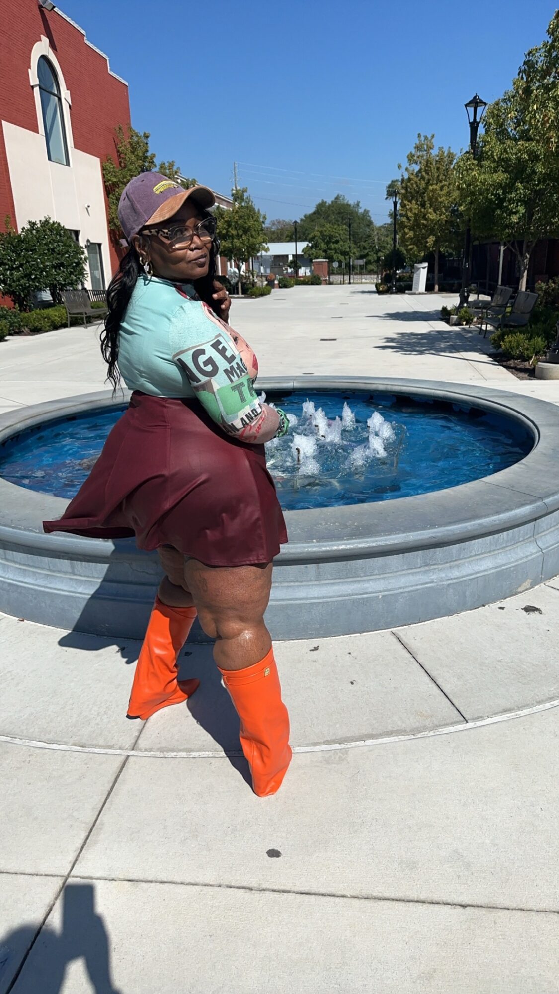 Person standing near a fountain in a town square, wearing a hat, glasses, a patterned shirt, a maroon skirt, and orange boots.