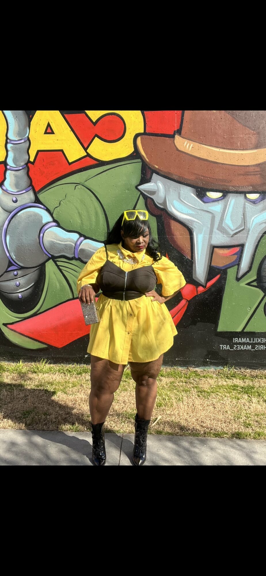 Person in yellow dress and black accessories standing in front of a colorful robot-themed mural.