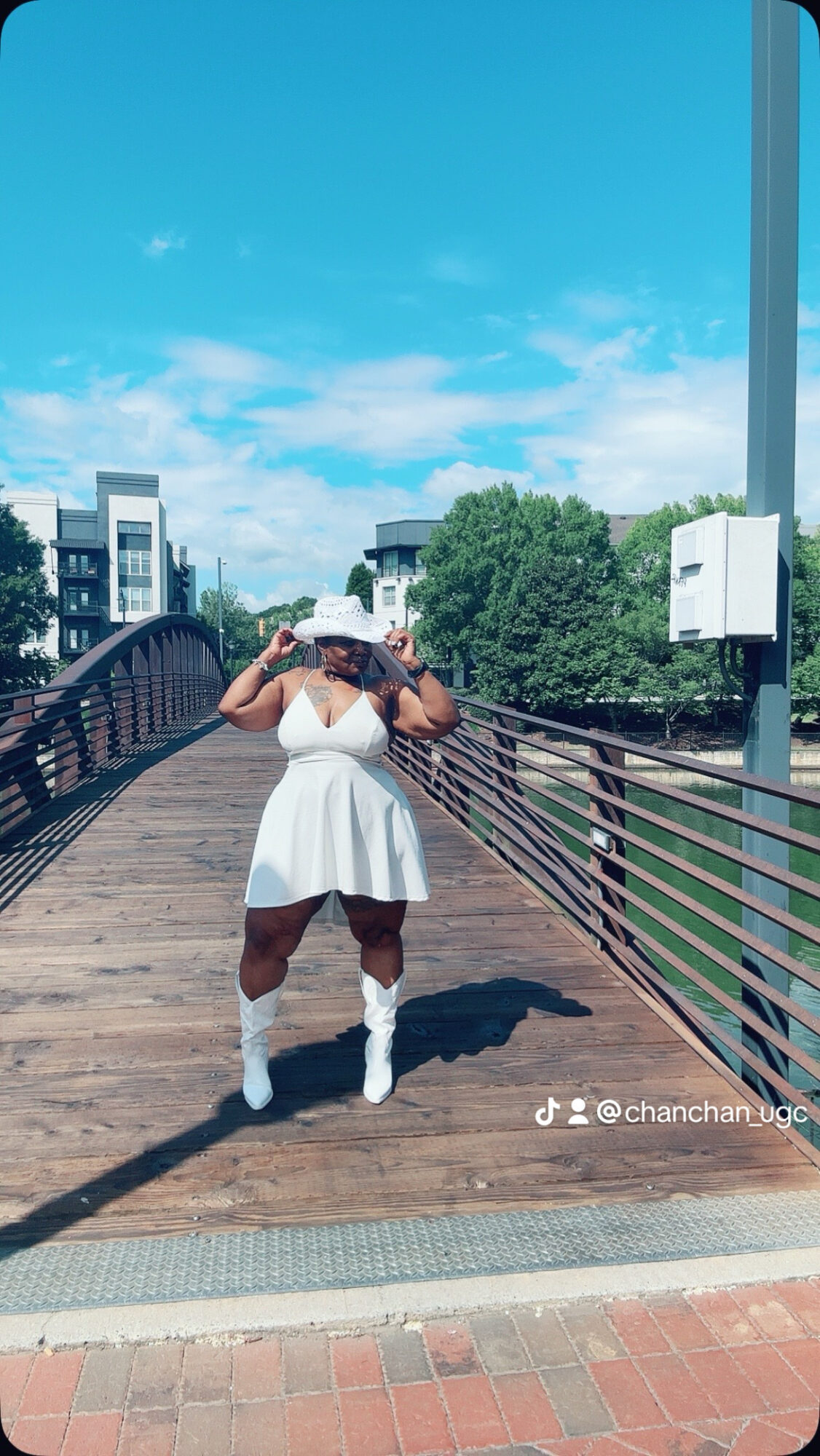 Person in white dress and hat standing on a wooden bridge with trees and buildings in background.