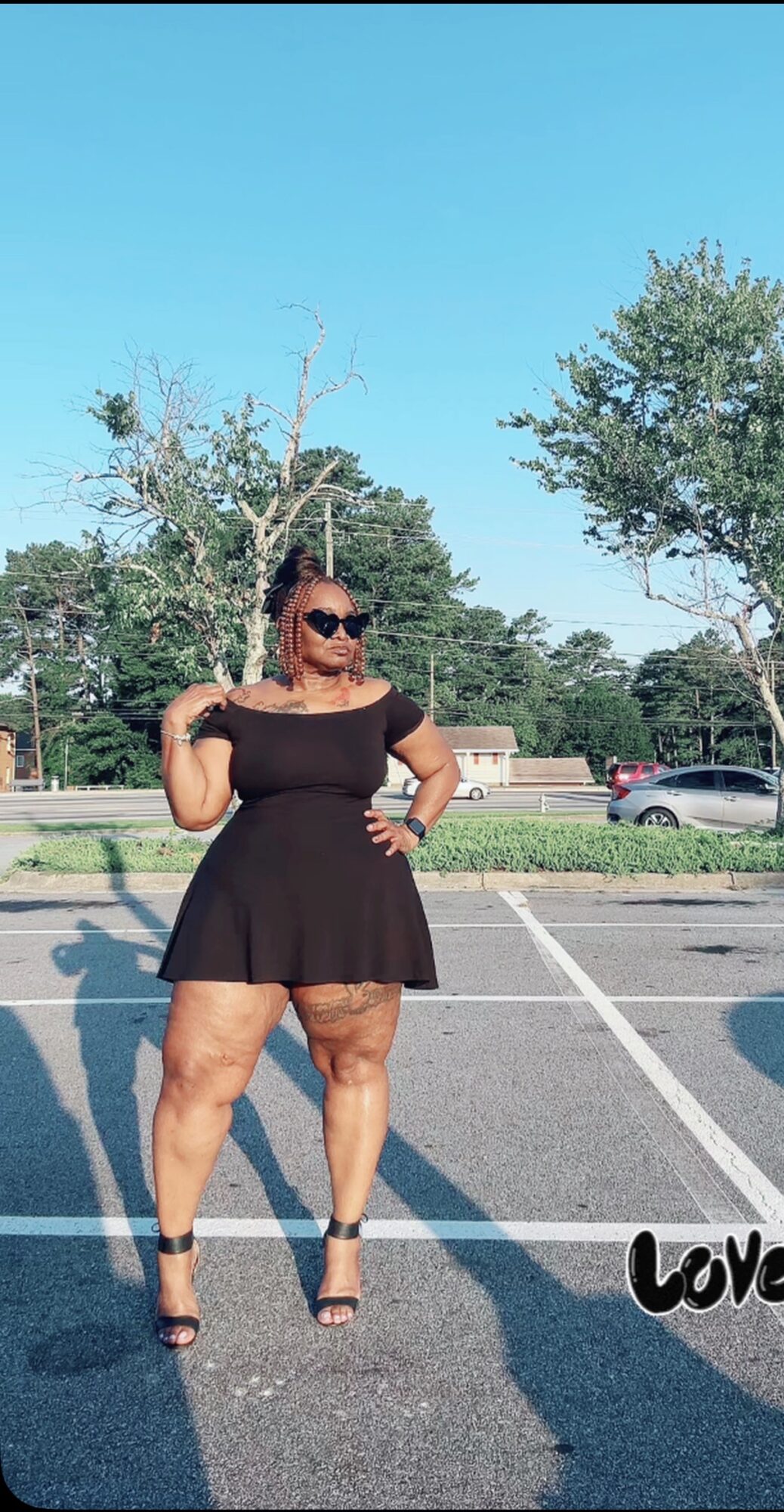Woman in black dress and sunglasses standing in parking lot with trees and cars in background.