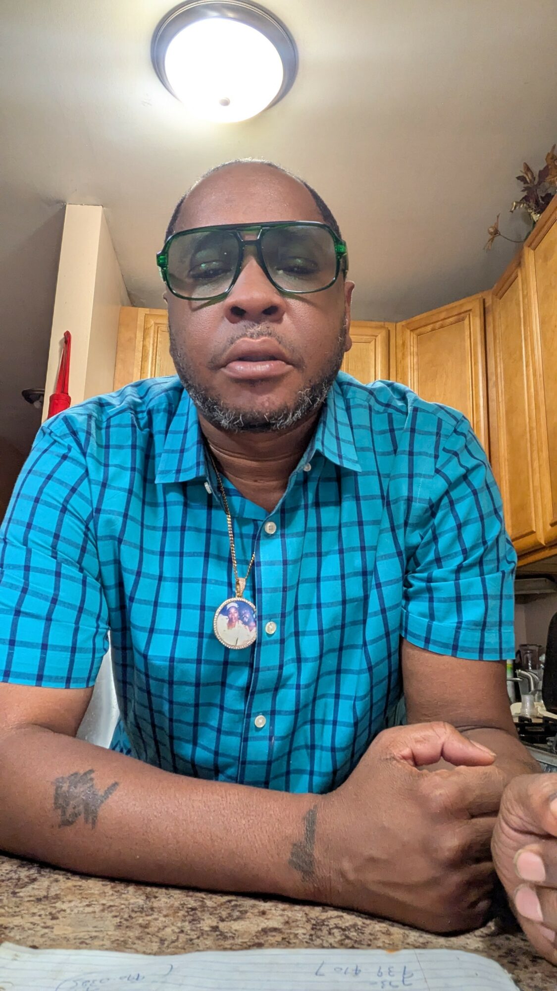 Man wearing glasses and a blue checkered shirt sitting at a kitchen counter with wooden cabinets in the background.