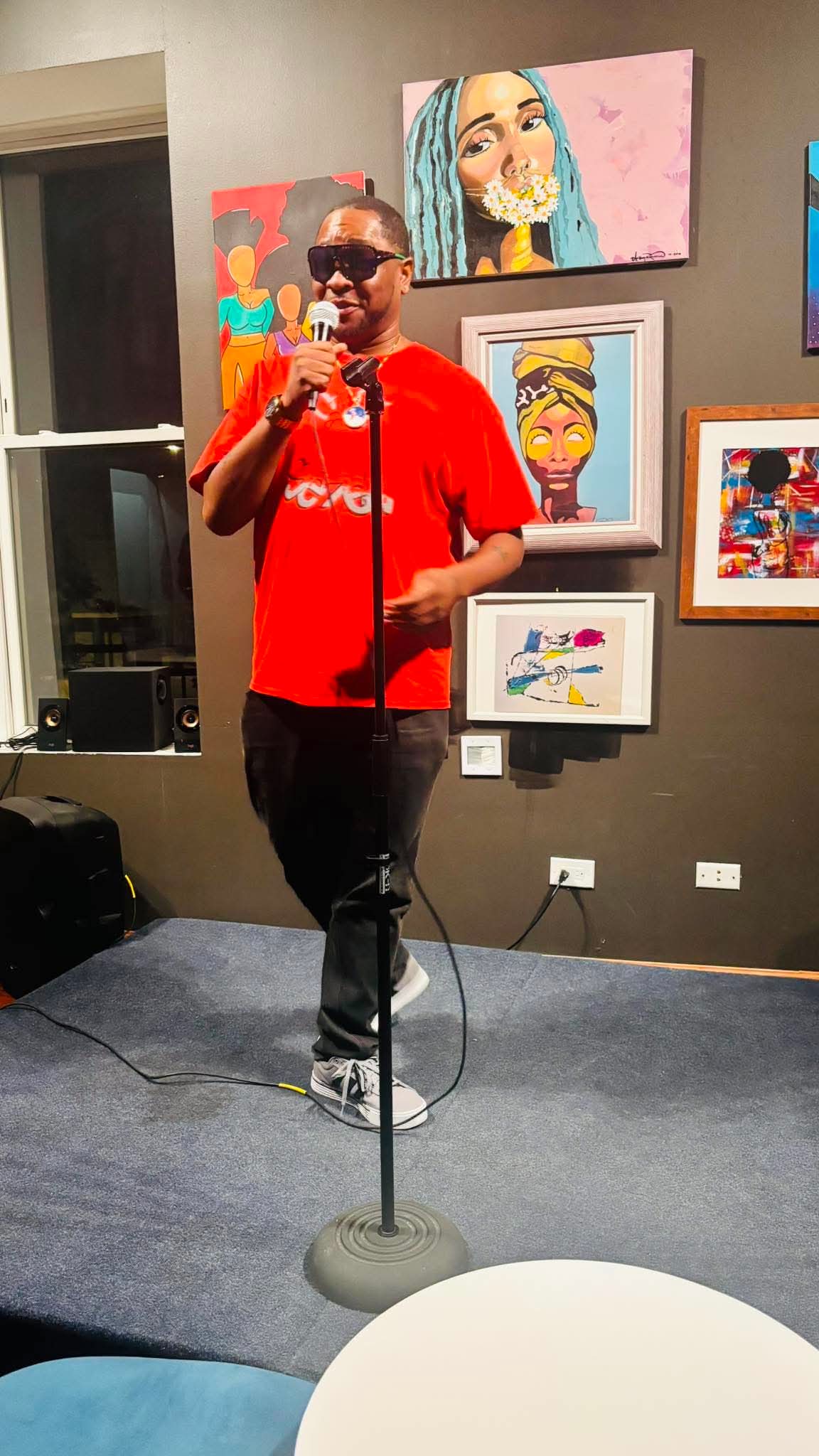 Person in red shirt speaking into microphone in art gallery with colorful paintings on wall.