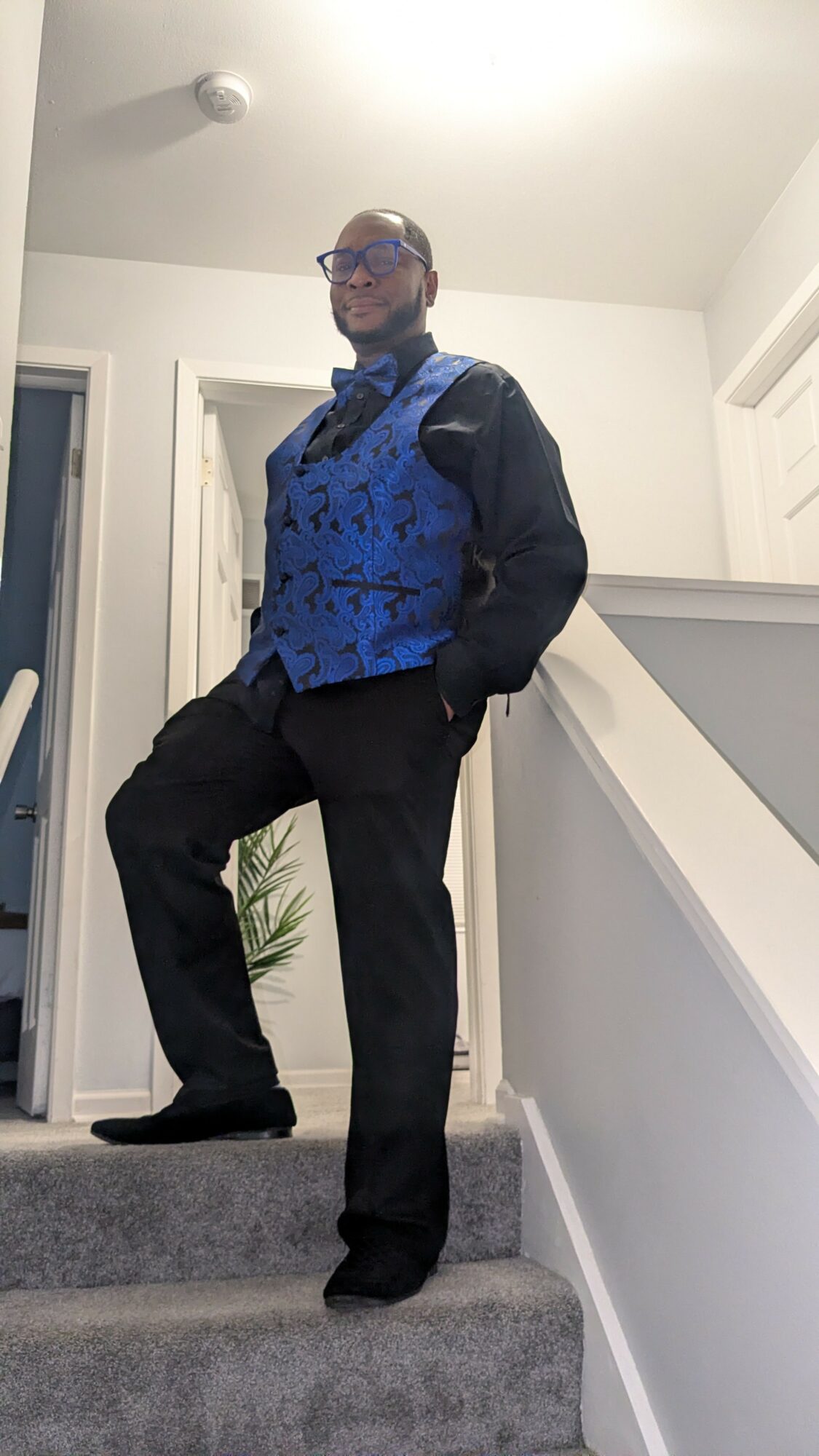 Person standing on stairs in a blue patterned vest, black jacket, and black pants, with hands in pockets, in a home interior.