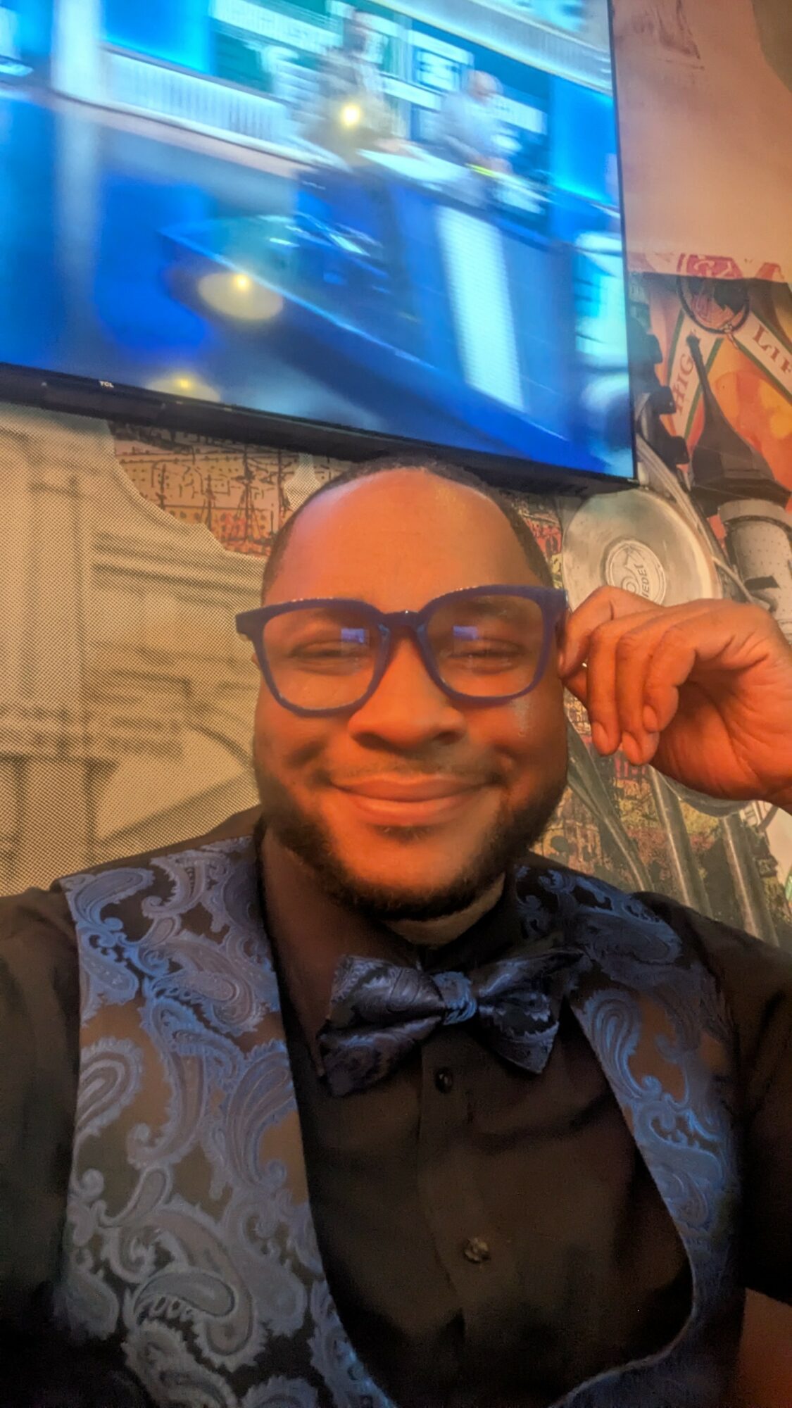 Person with glasses smiling, wearing a dark shirt and patterned vest, sitting indoors with a TV and wall art behind.