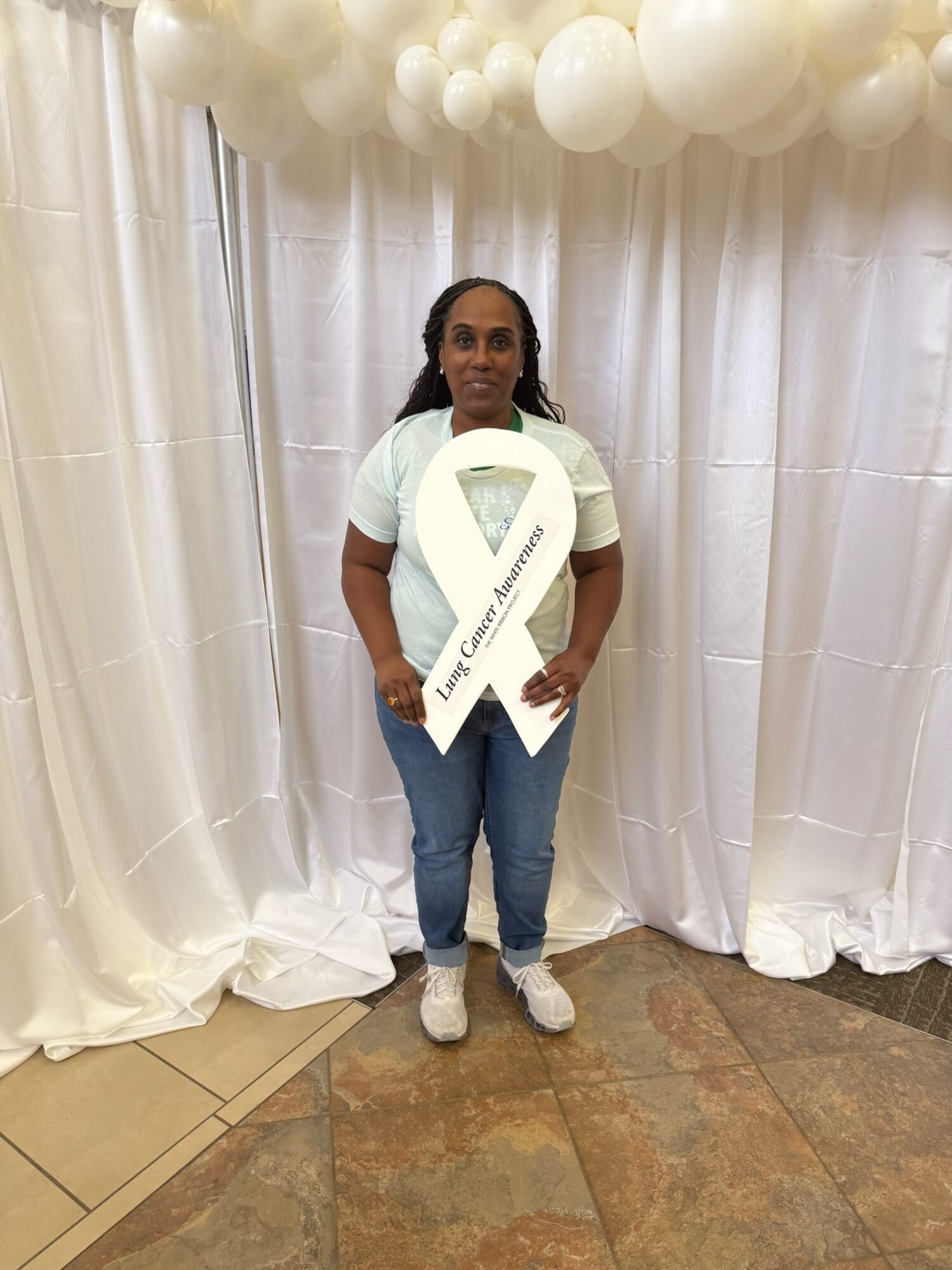 Woman standing in front of white curtain with balloons, holding a large white key-shaped sign, wearing a white shirt and jeans.