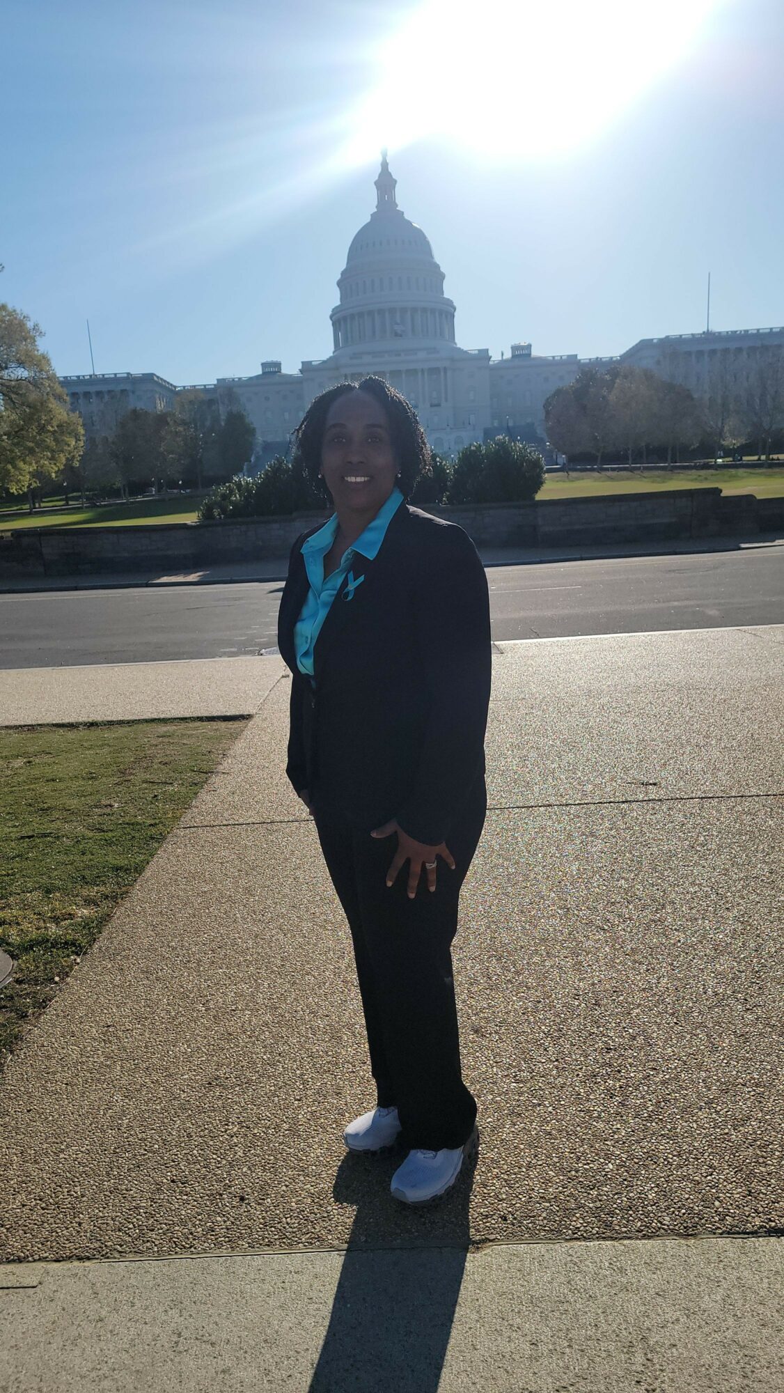 Person standing outdoors on a sidewalk with a cityscape in the background, sunlight shining brightly.