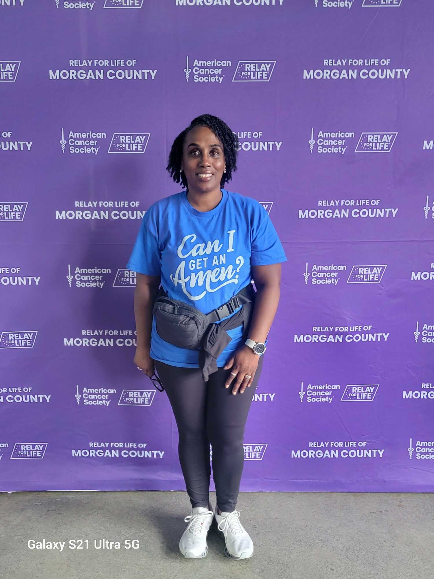 Woman standing in front of purple backdrop with white text, wearing a blue shirt, black pants, and white shoes.
