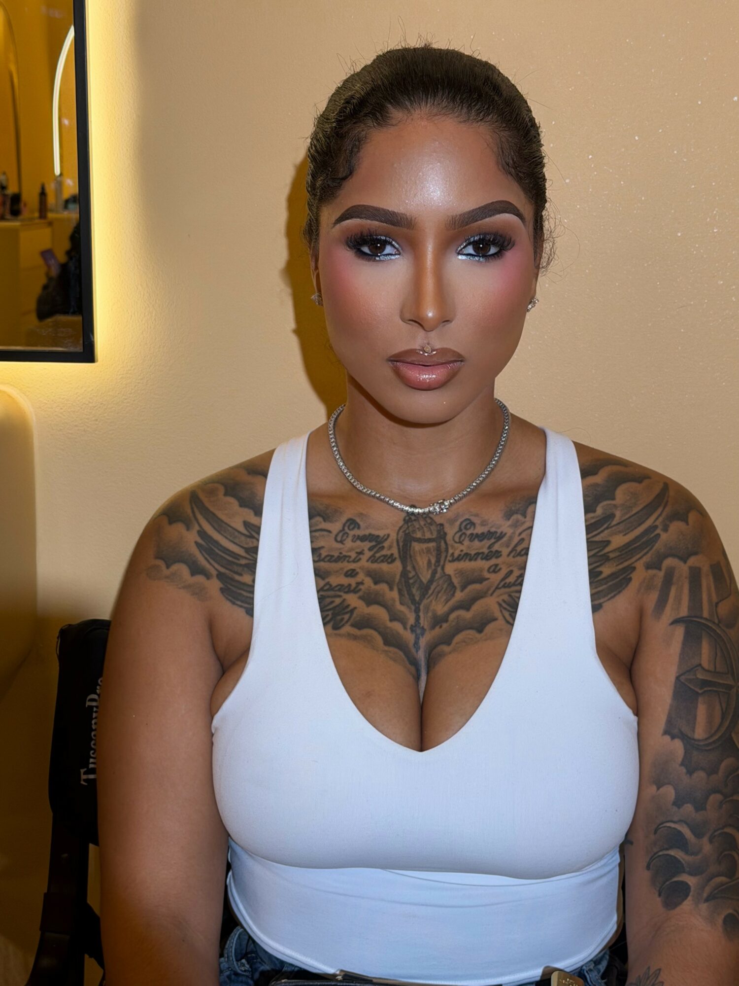 Woman with tattoos on arms and chest, wearing a white tank top and necklace, standing indoors against a beige wall.