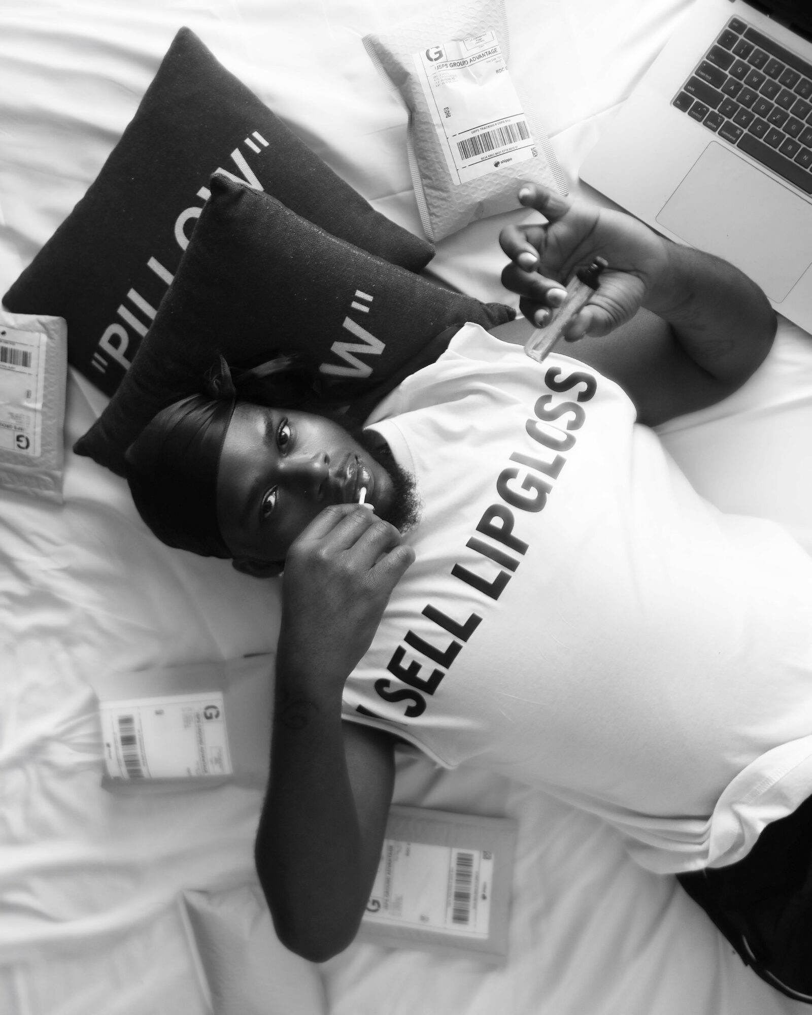Person lying on bed with pillows, surrounded by milk cartons, using a laptop, wearing a white shirt with black text.
