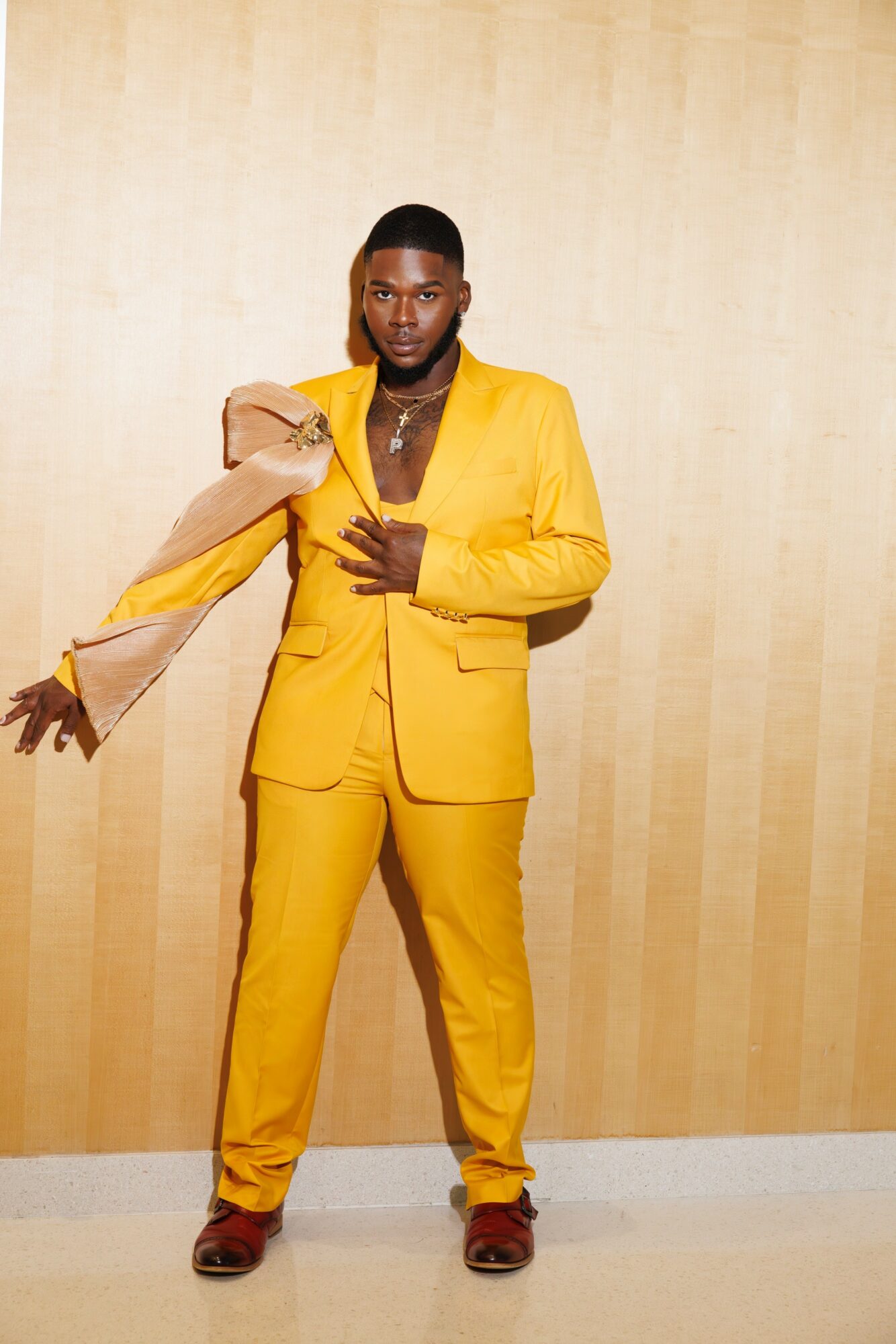 Person in a yellow suit holding a beige bag, standing against a beige wall, looking at the camera.