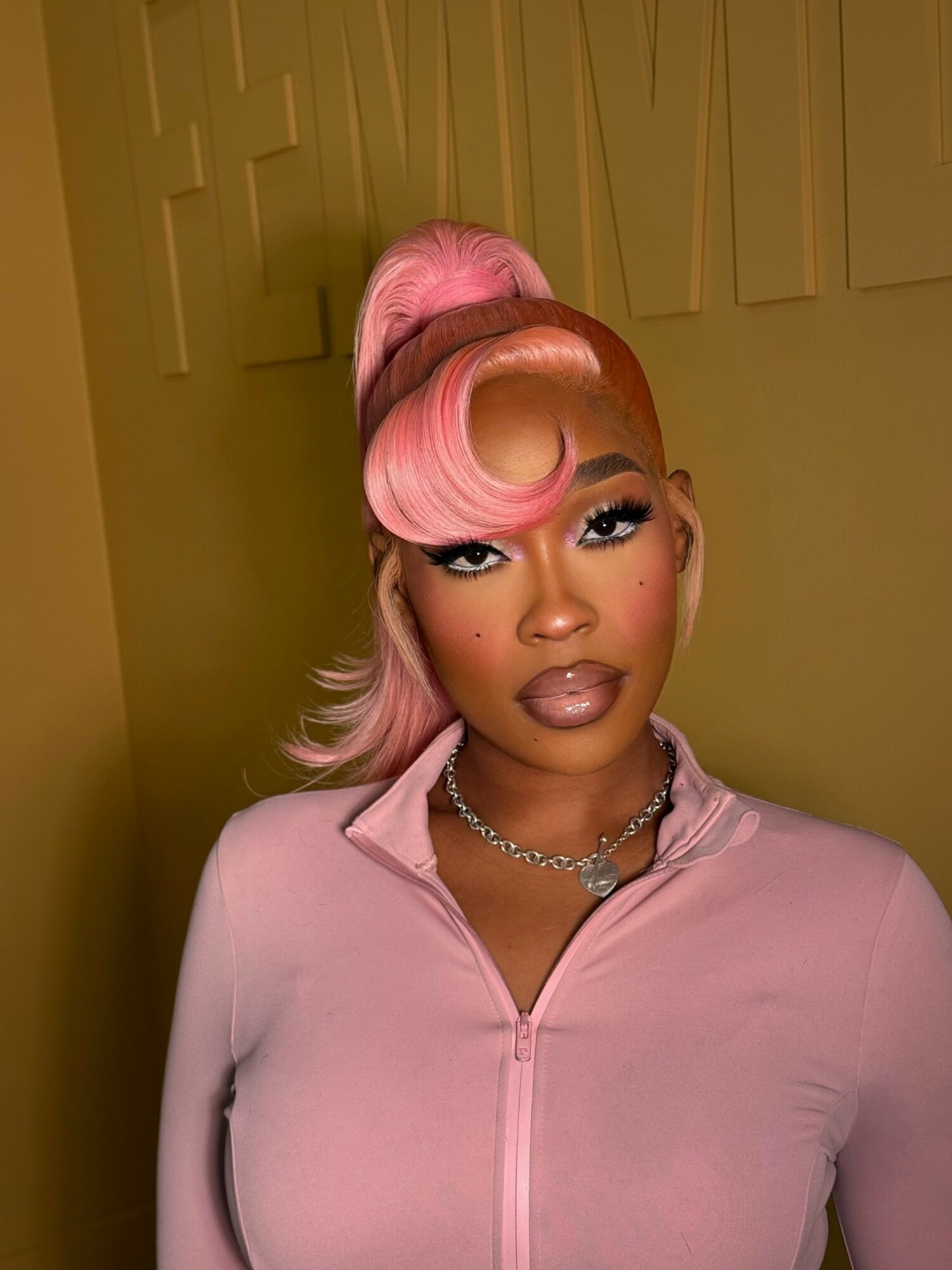 Woman with pink hair styled in an updo, wearing a light pink top and necklace, against a yellow wall.