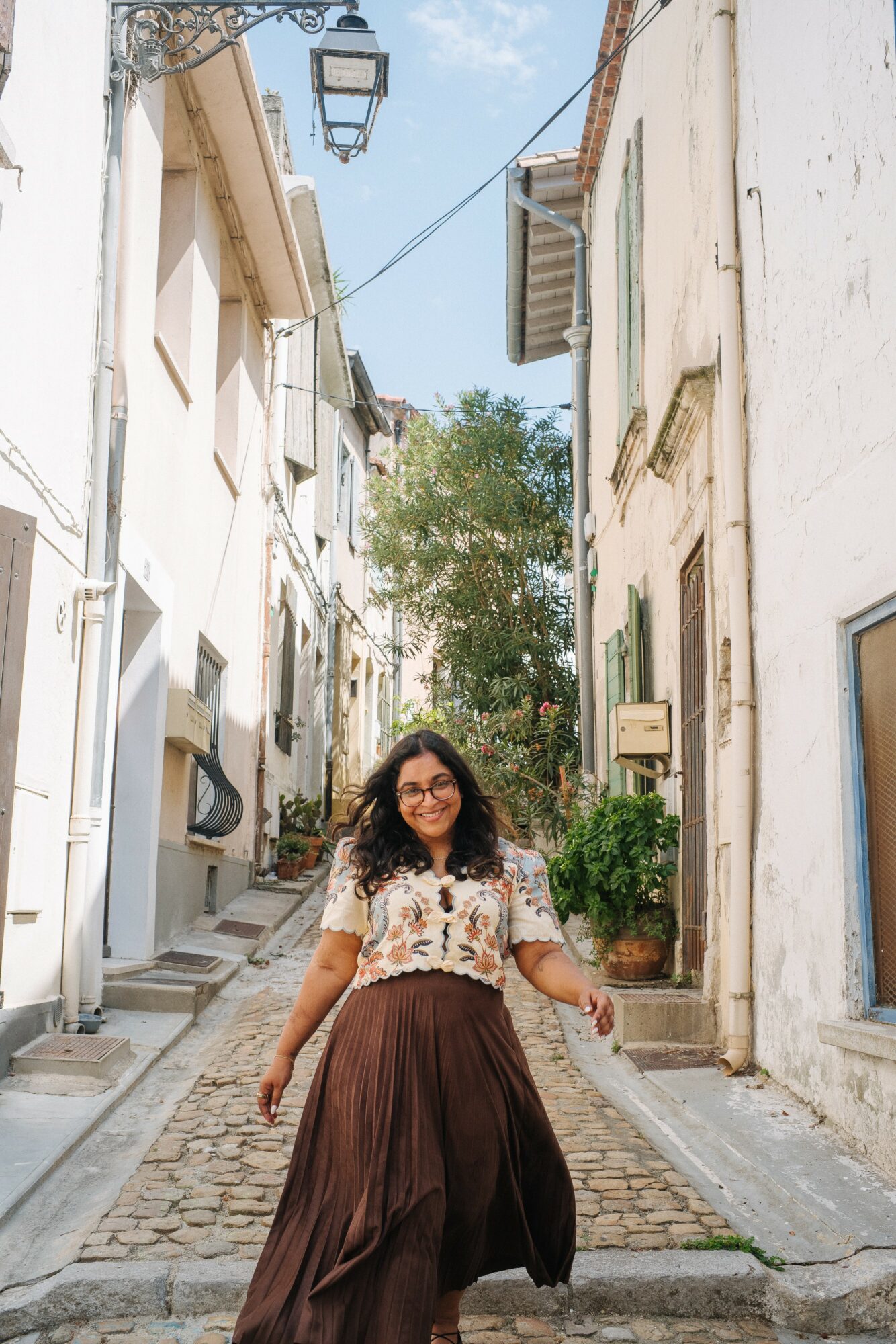 Young woman with glasses and curly hair standing on a narrow cobblestone street, smiling, with white buildings and greenery around.
