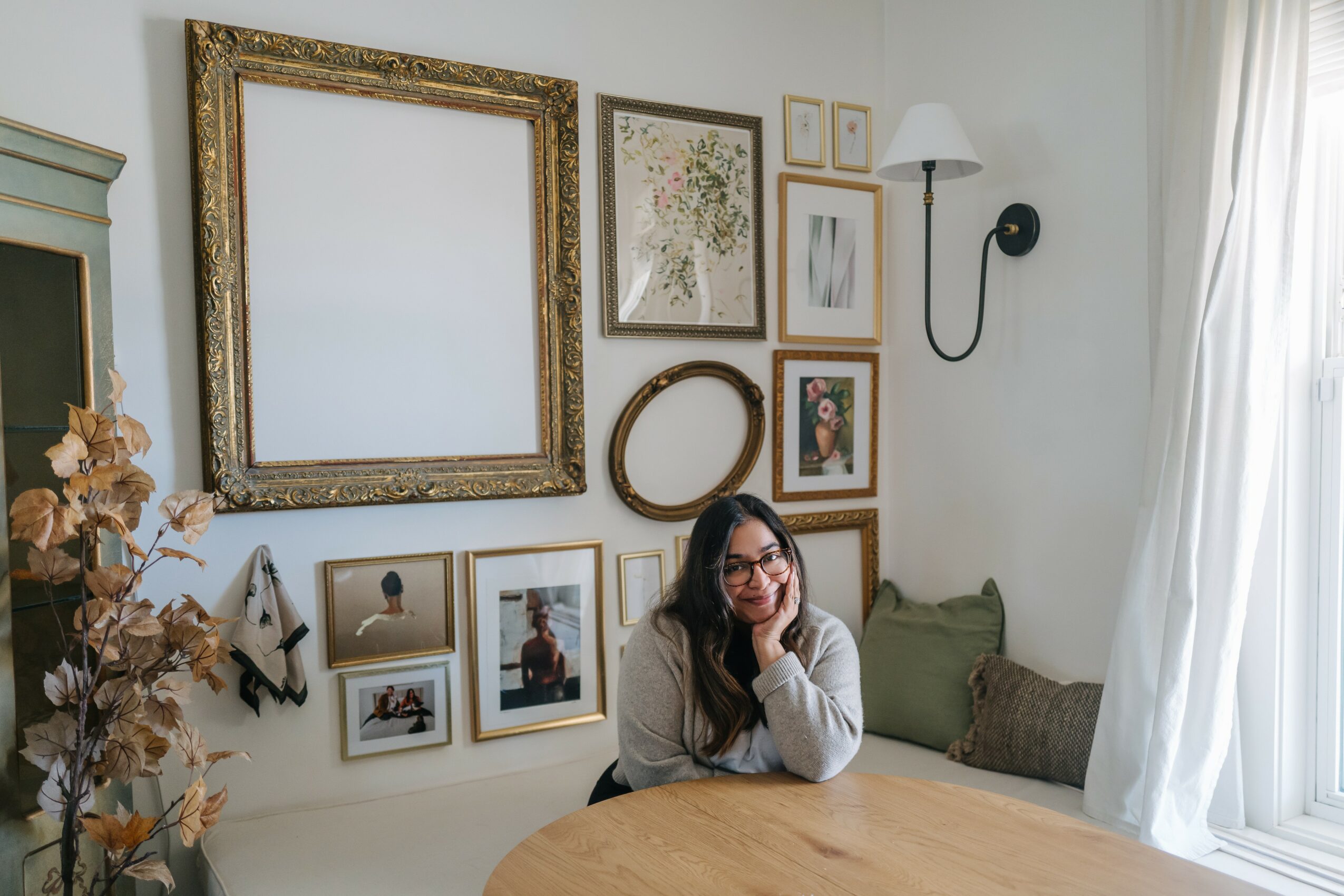 Woman with glasses sitting at a wooden table in a decorated room with framed pictures and a window with curtains.