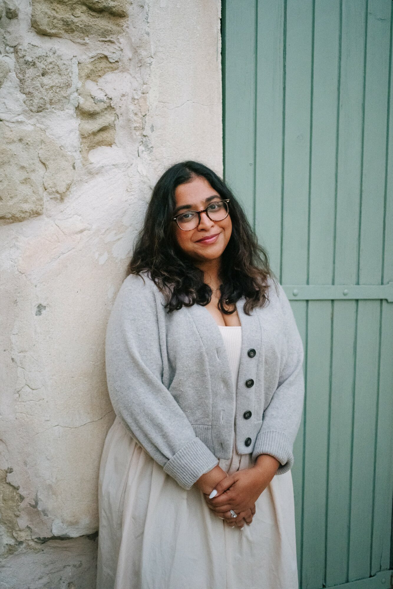 Woman with glasses and dark hair, wearing a light cardigan and dress, standing outdoors near a stone wall and green door.