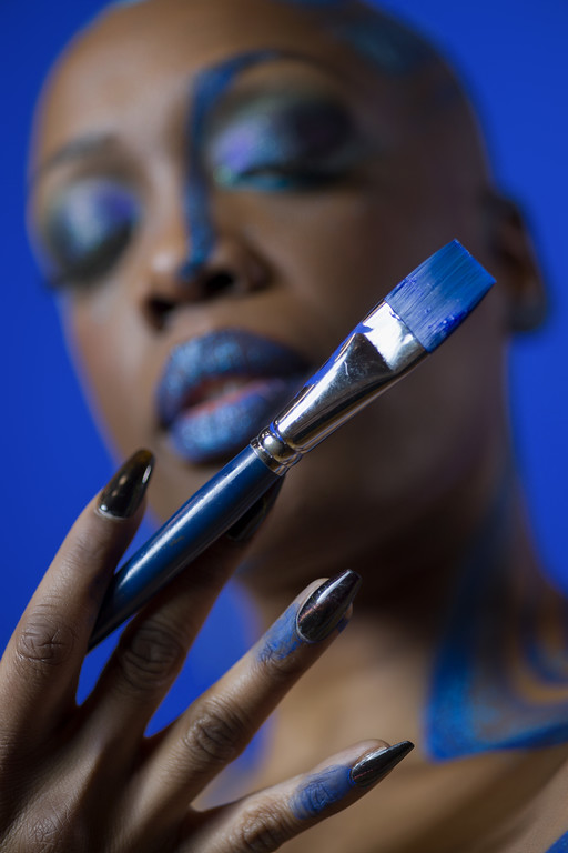 Close-up of a person with vibrant blue makeup holding a paintbrush near their face, against a blue background.