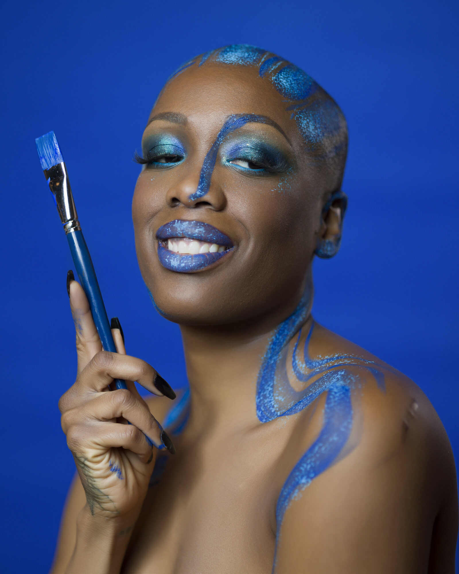 Woman with blue makeup and paint, holding a paintbrush, smiling against a blue background.