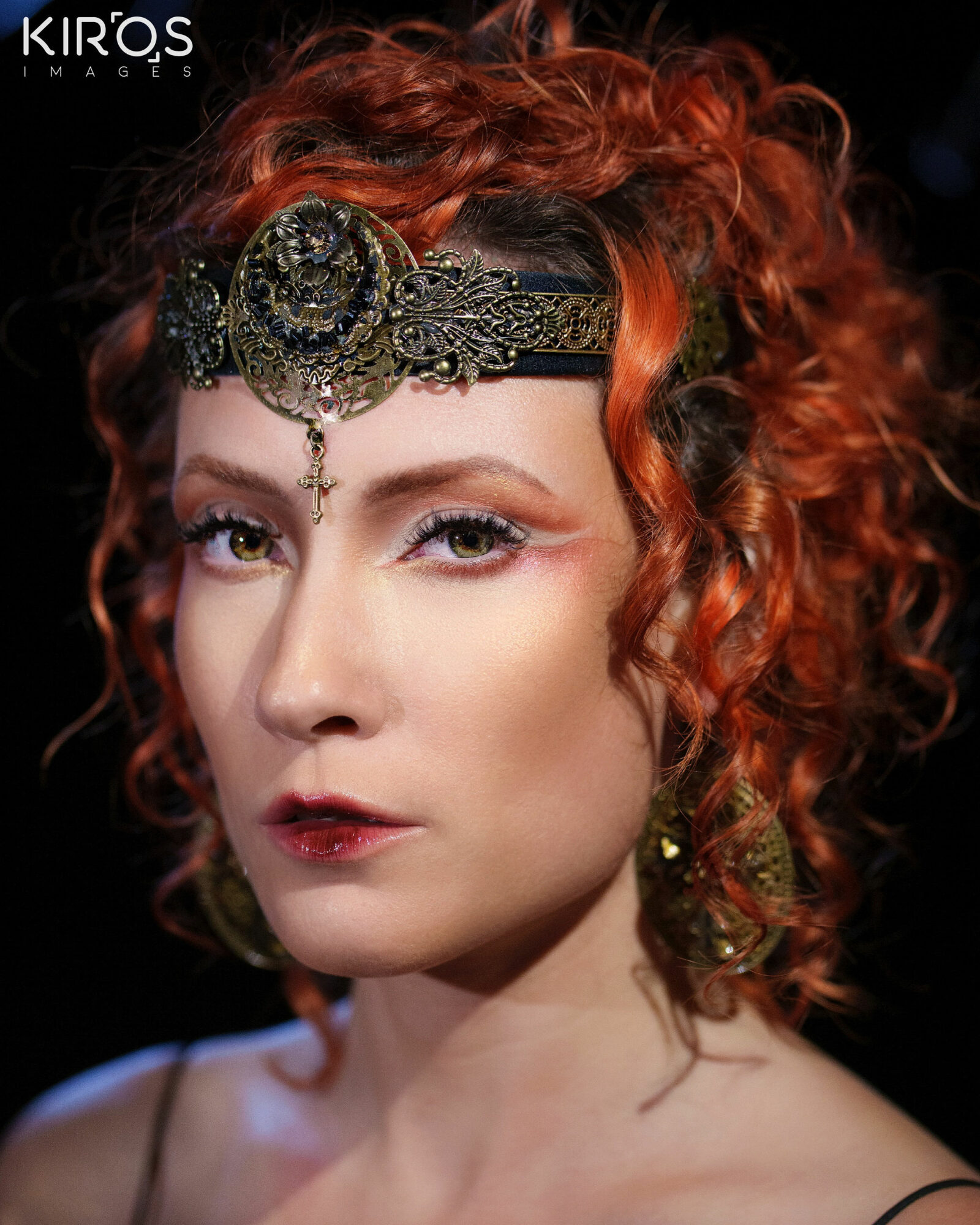 Woman with curly red hair wearing ornate headpiece and earrings, looking at camera against dark background.