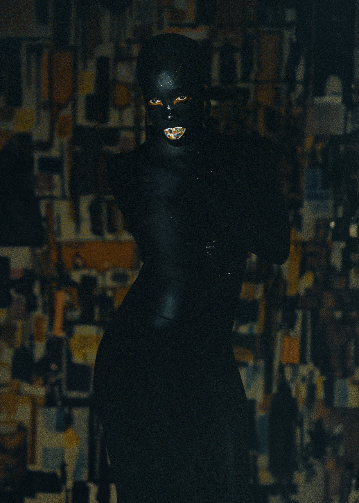 Person with dark skin and bright eyes, smiling, in a dark setting with a patterned background.