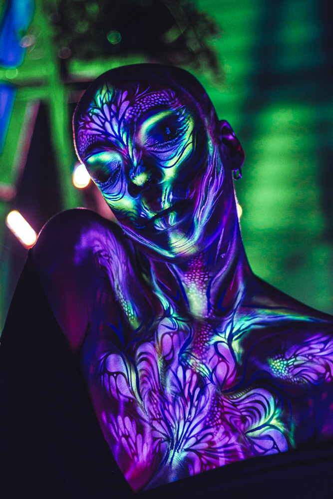 Person with glowing, colorful body paint and face, illuminated under blacklight, with a dark background.