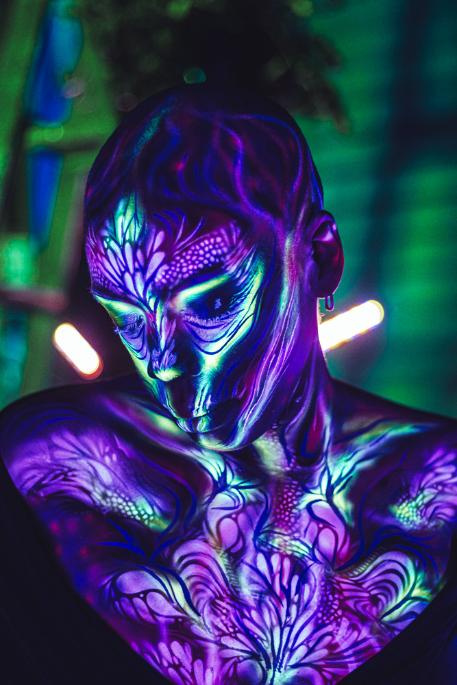 Person with glowing, intricate body paint and face design under blacklight, in a dark environment with colorful lights.