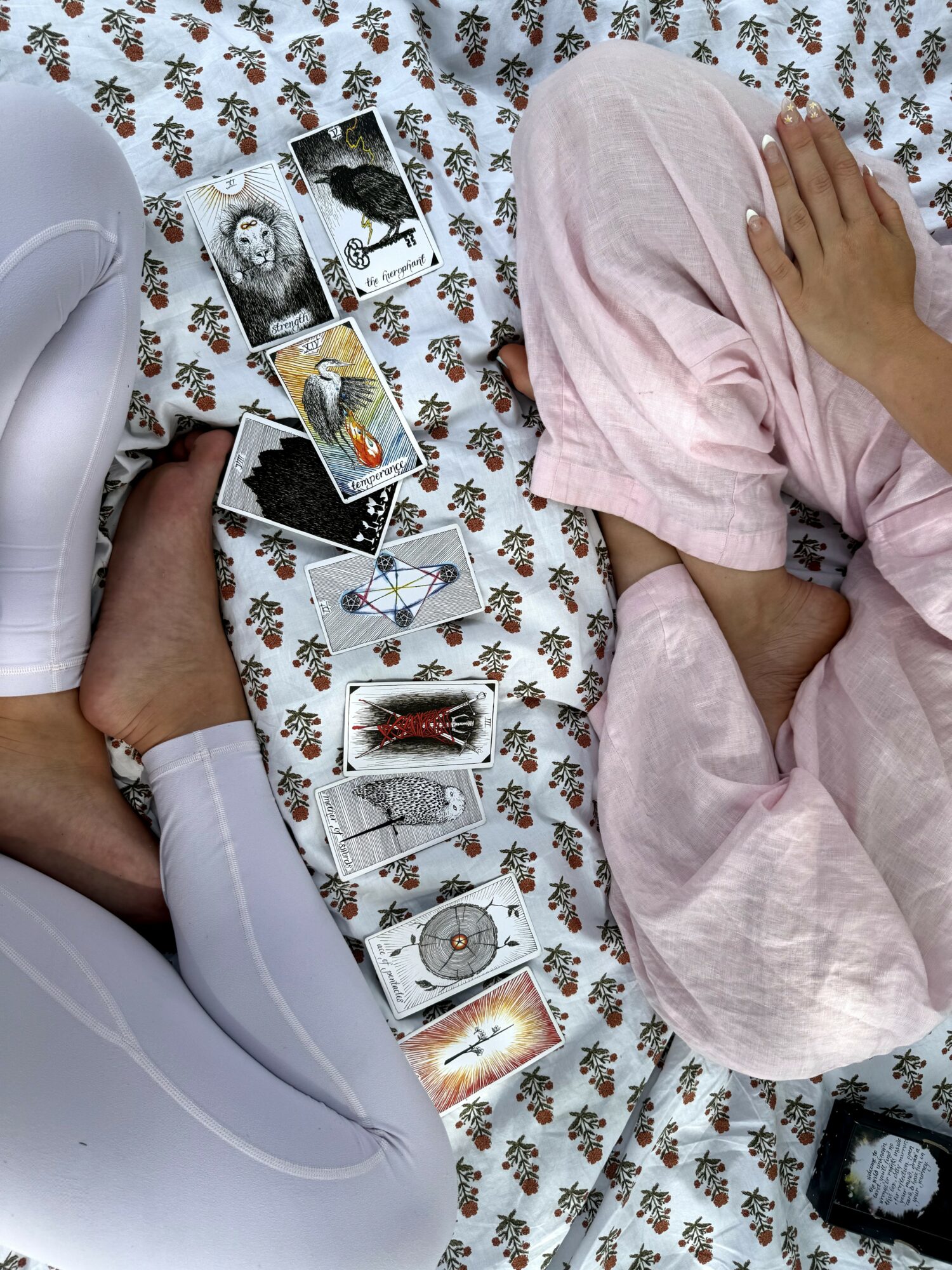 Two people sitting on a floral bedspread with tarot cards and photographs spread between them, one person wearing white pants and the other pink pants.