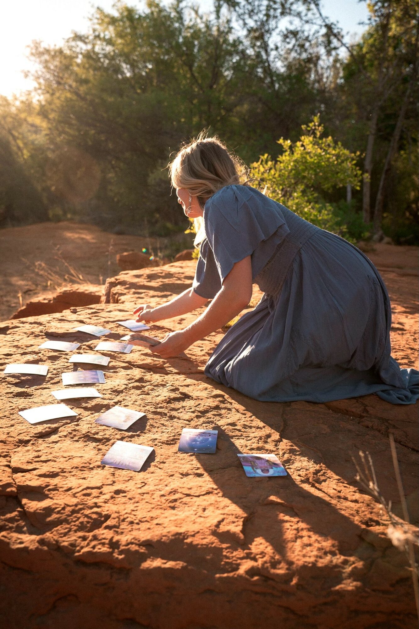 Girl in a blue dress kneeling outdoors, arranging small objects on a dirt surface, with trees and sunlight in the background.