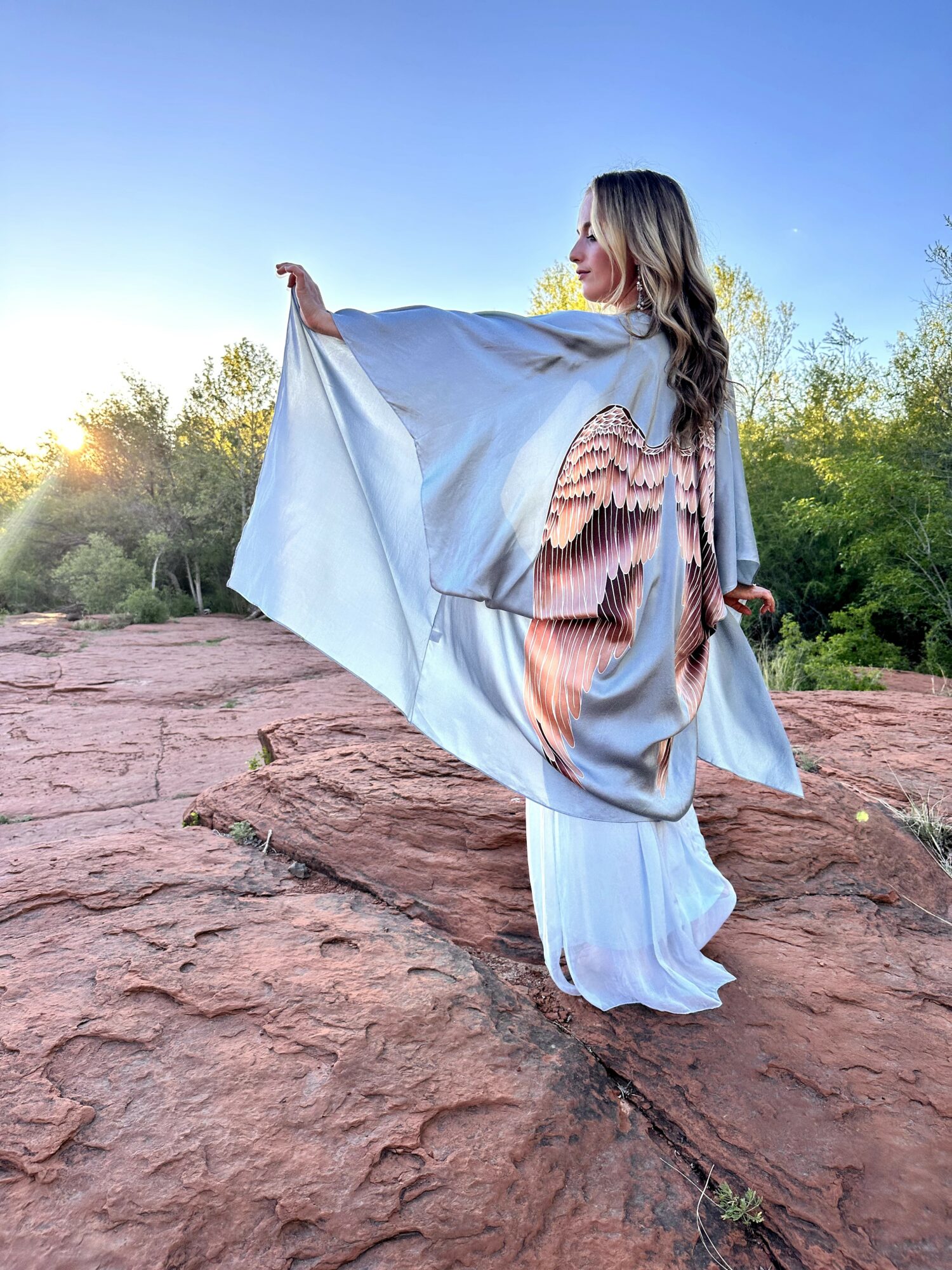 Woman standing on rocky terrain holding a large, flowing fabric outdoors with trees and a clear sky in the background.