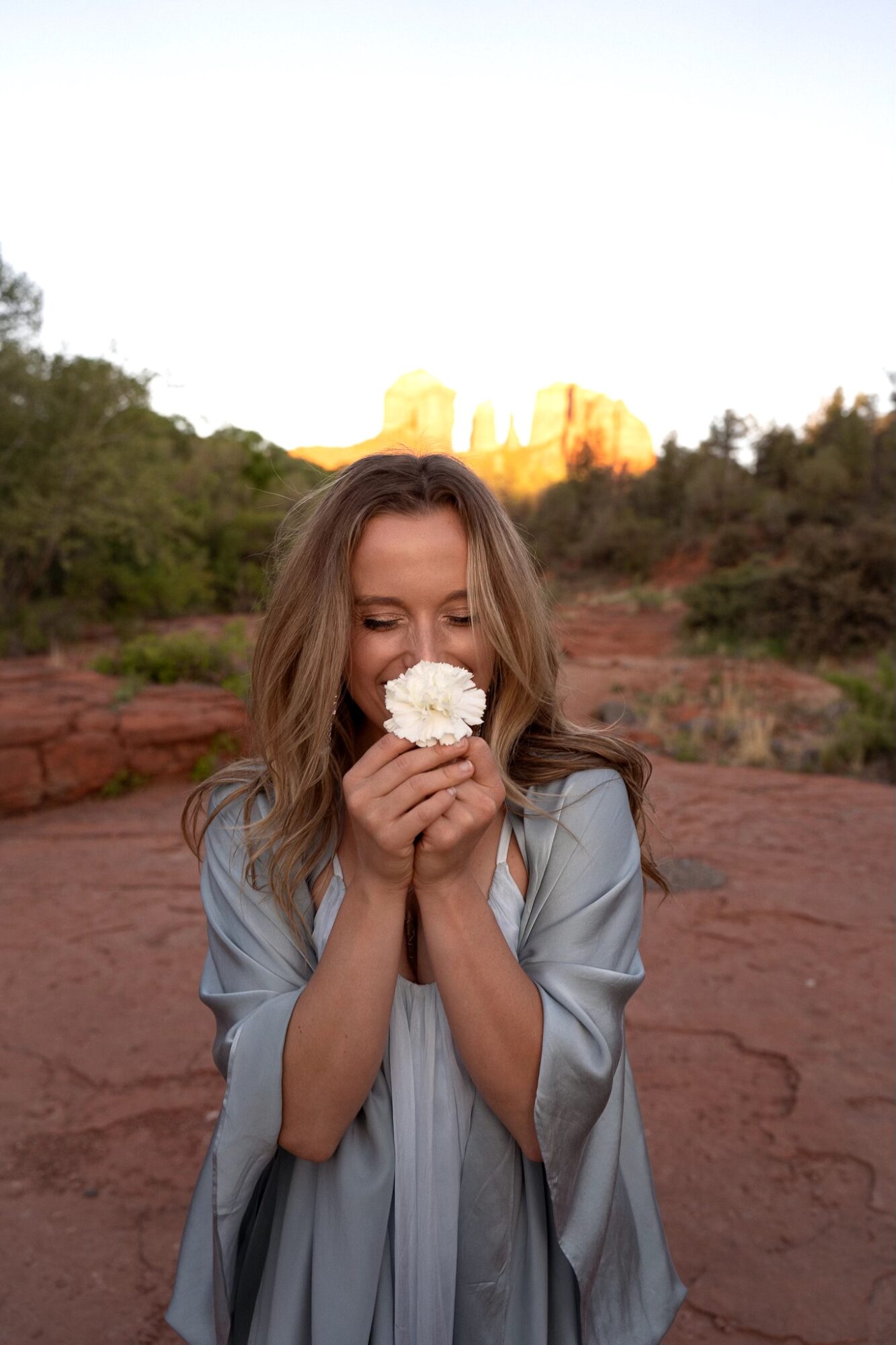 Woman holding a flower close to her face outdoors with a landscape and sky in the background.