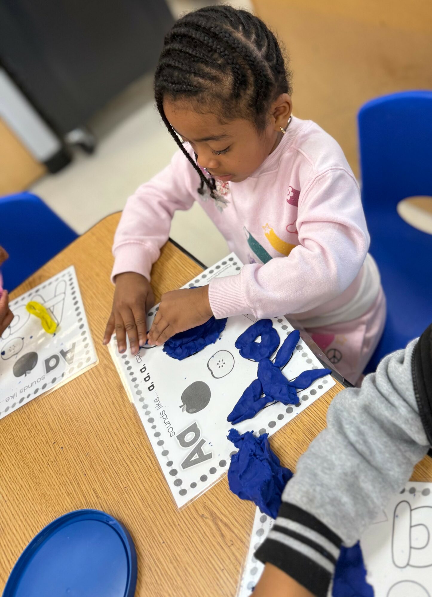 Young girl with braided hair working on a craft activity at a table, with a blue chair nearby.