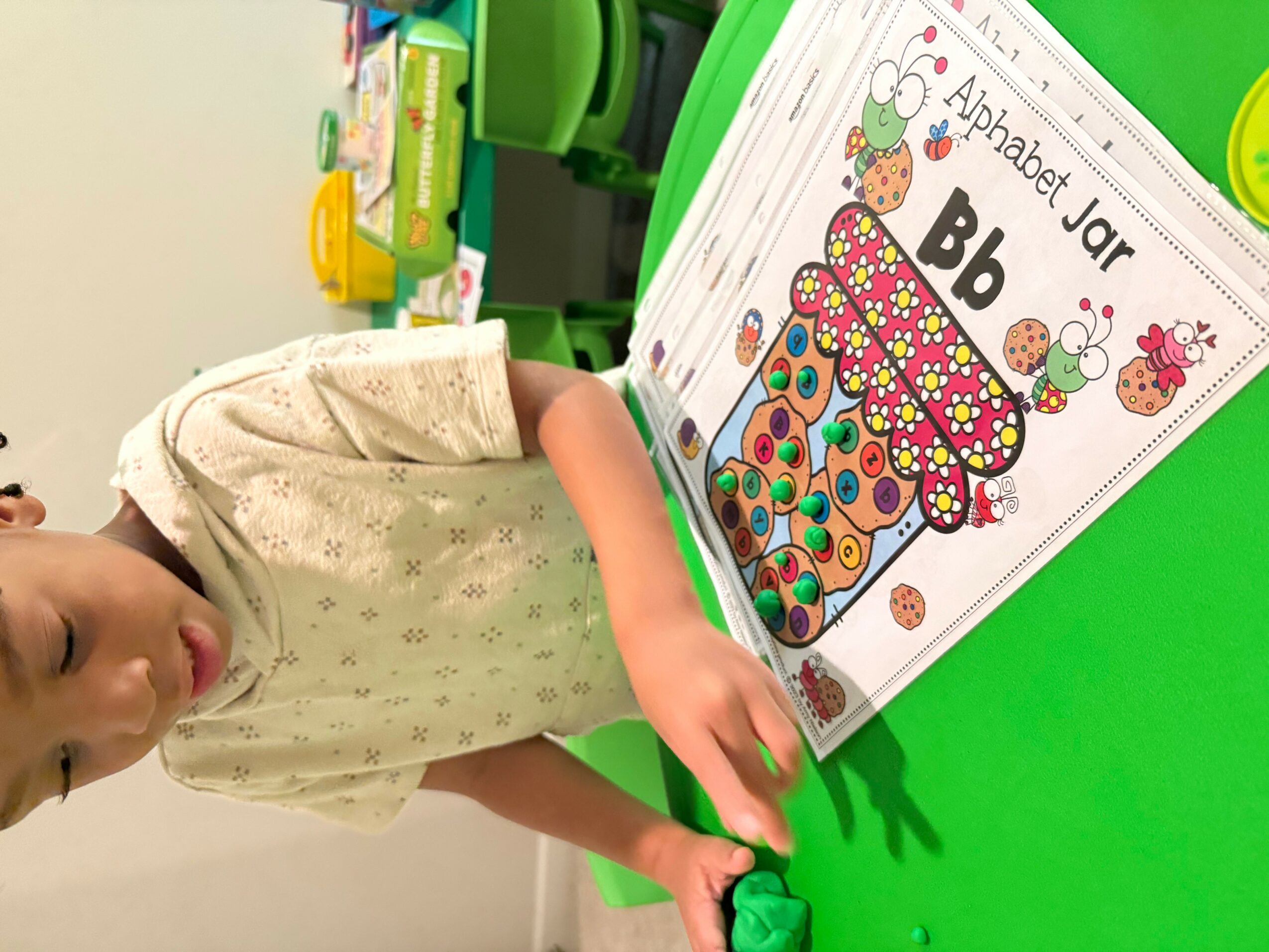 Child sitting at a green table with an alphabet jar coloring page, holding a green crayon, in a classroom.