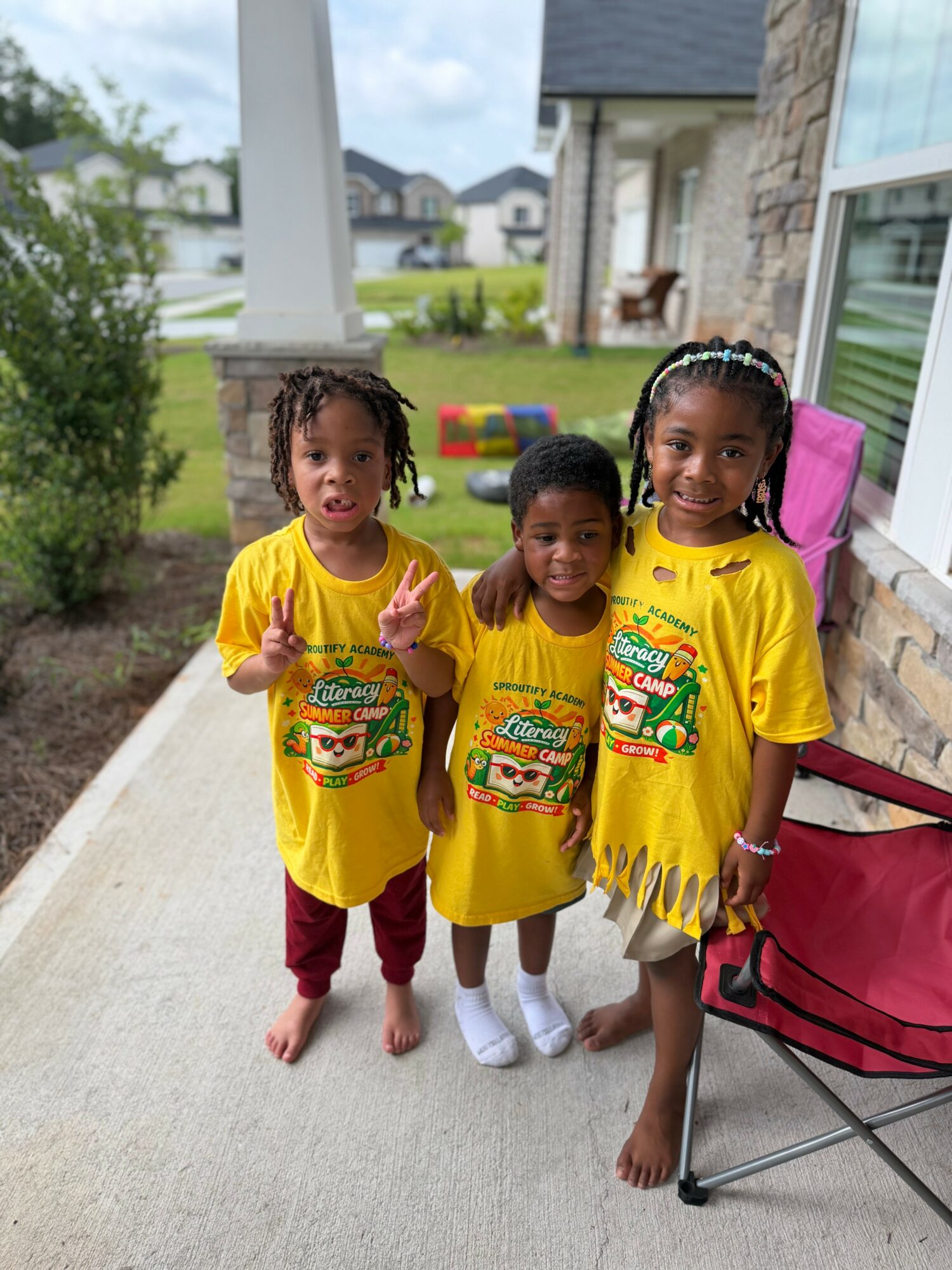 Three children in yellow shirts standing outside on a sidewalk, smiling, with houses and greenery in the background.