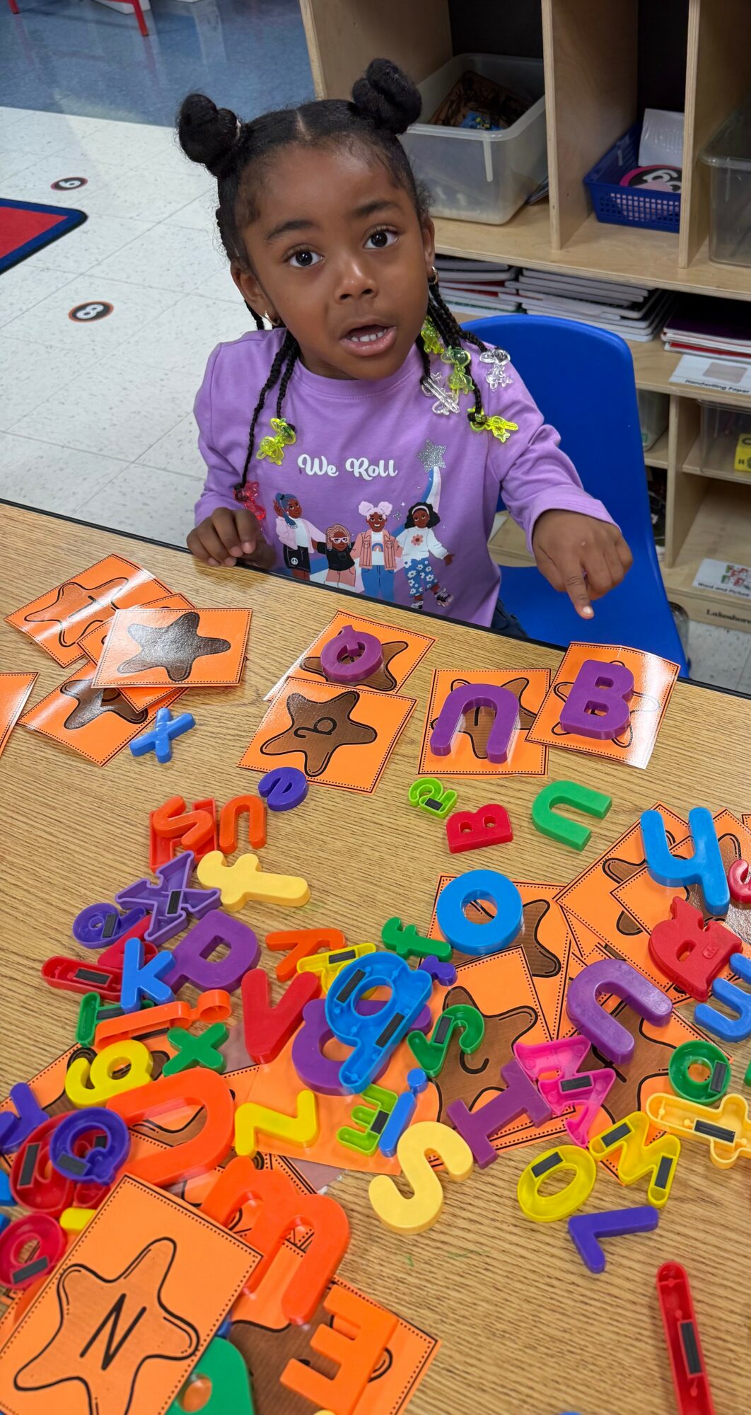 Young girl with braided hair and purple shirt sitting at a table with colorful foam letter puzzles and foam letters scattered around.